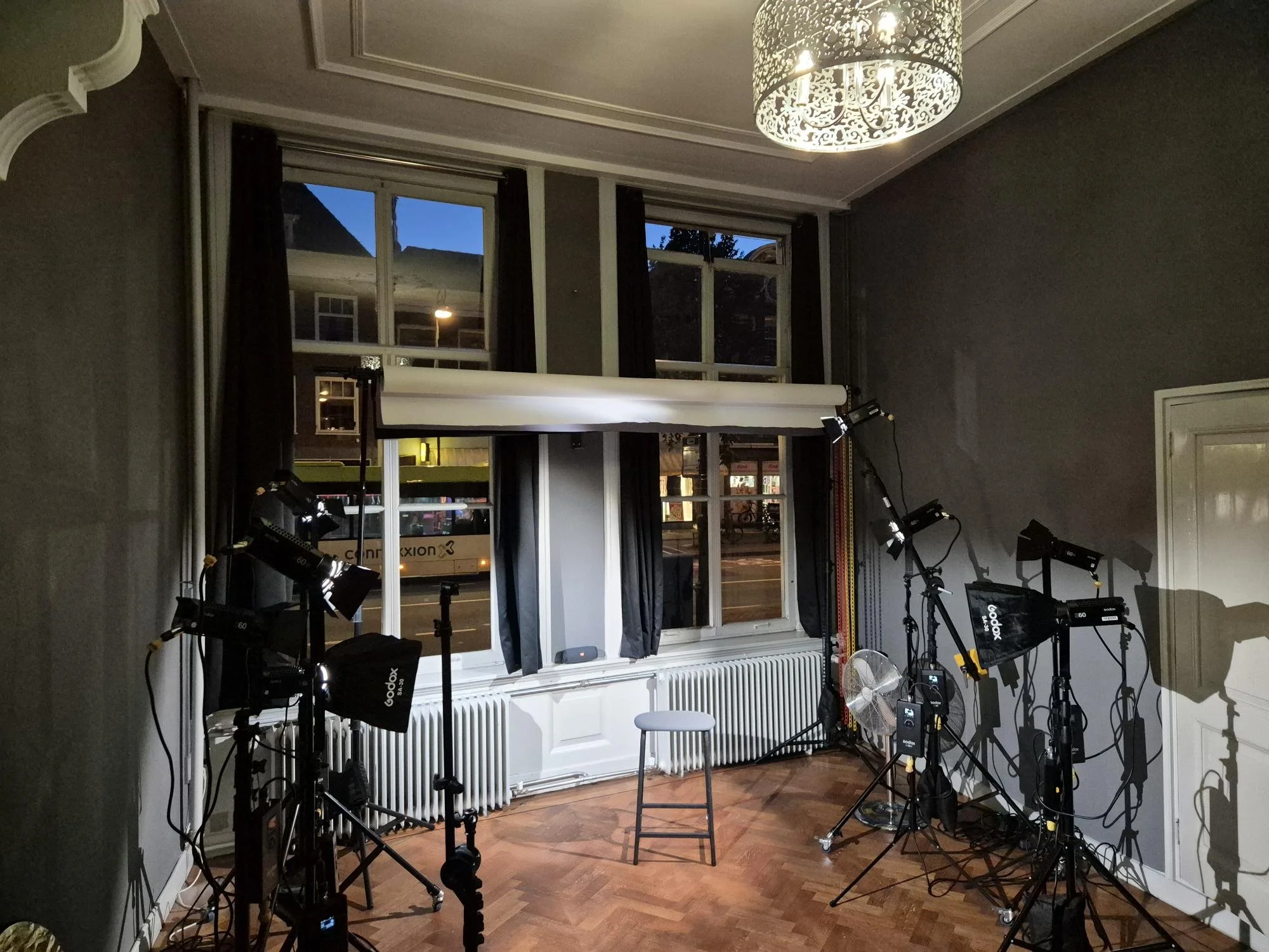Binnenruimte met grote ramen, diverse professionele film- of fotografieapparatuur, zoals lampen, statieven, en een stoel, met een verlichtingsarmatuur aan het plafond. Buiten is het donker, zichtbaar door de ramen. De ruimte heeft houten vloer en witte radiatoren onder de ramen.