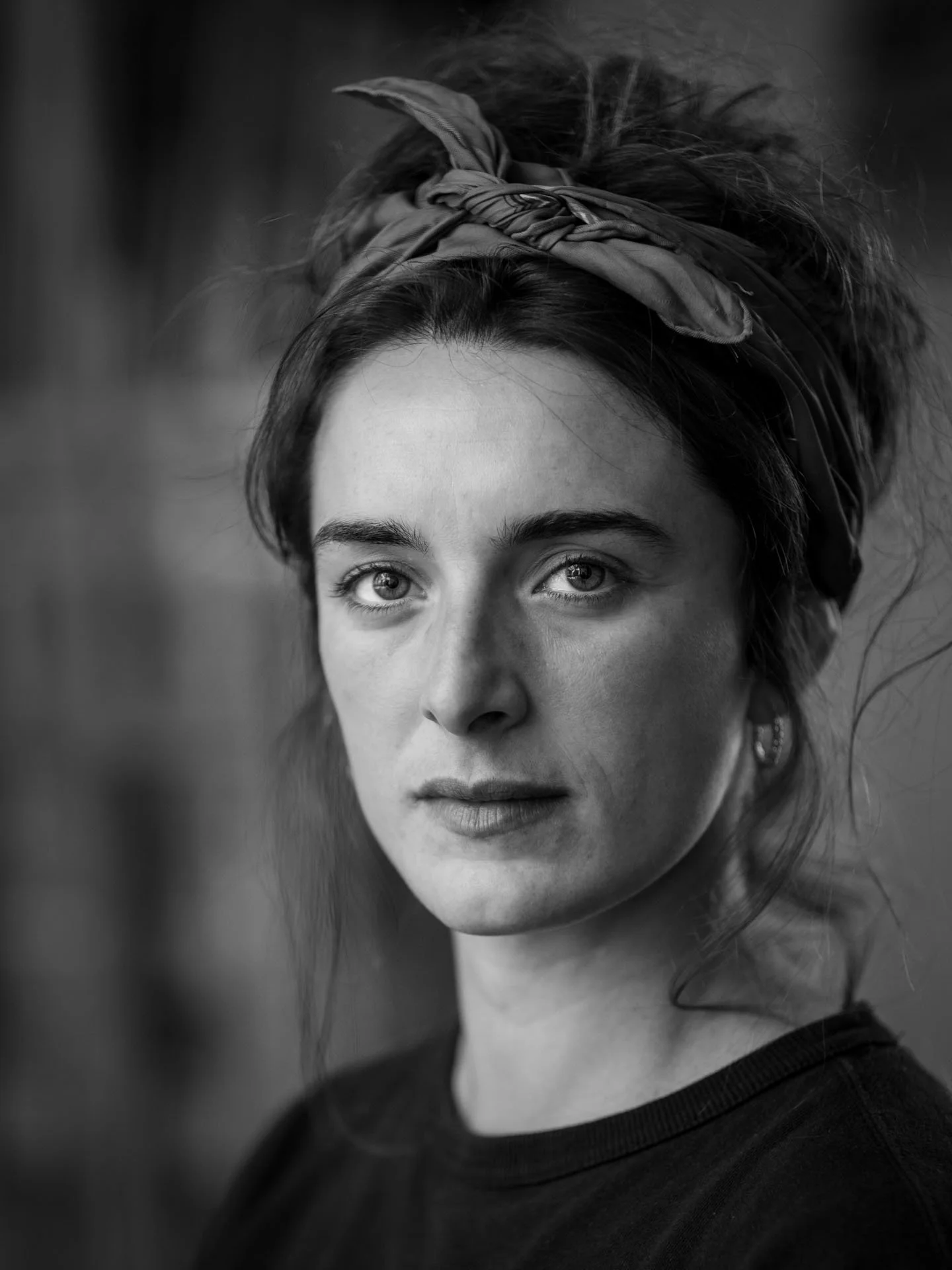 Zwarte en witte portretfoto van een jonge vrouw met een haarband, naar links kijkend, met een donkere achtergrond.