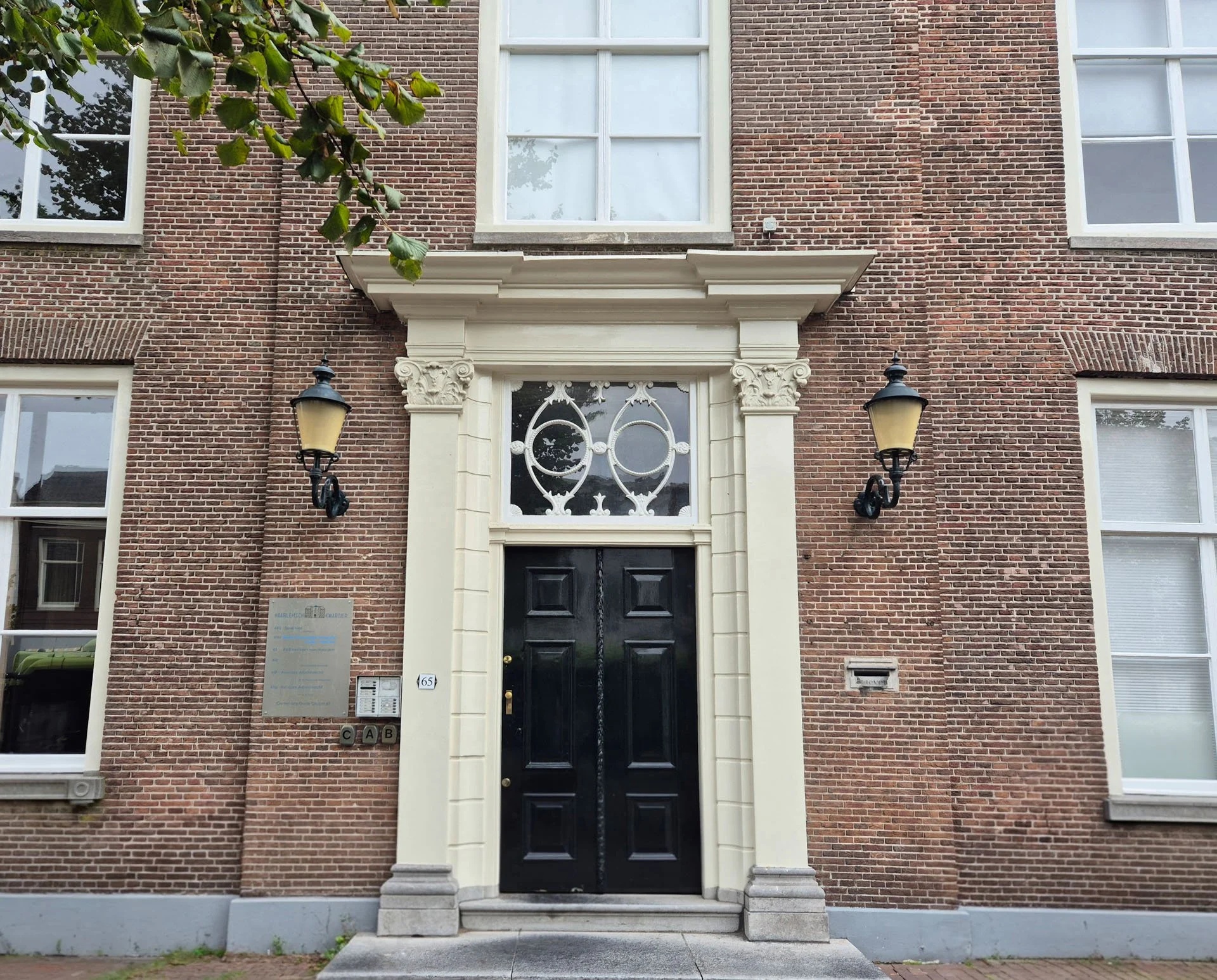 De gevel van een wit gerestaureerd rood bakstenen huis met een zwarte voordeur en witte omlijsting. Boven de deur is een decoratief wit lijstwerk met een klein uitbouw. Er hangen twee zwarte buitenlampen aan weerszijden van de deur.
