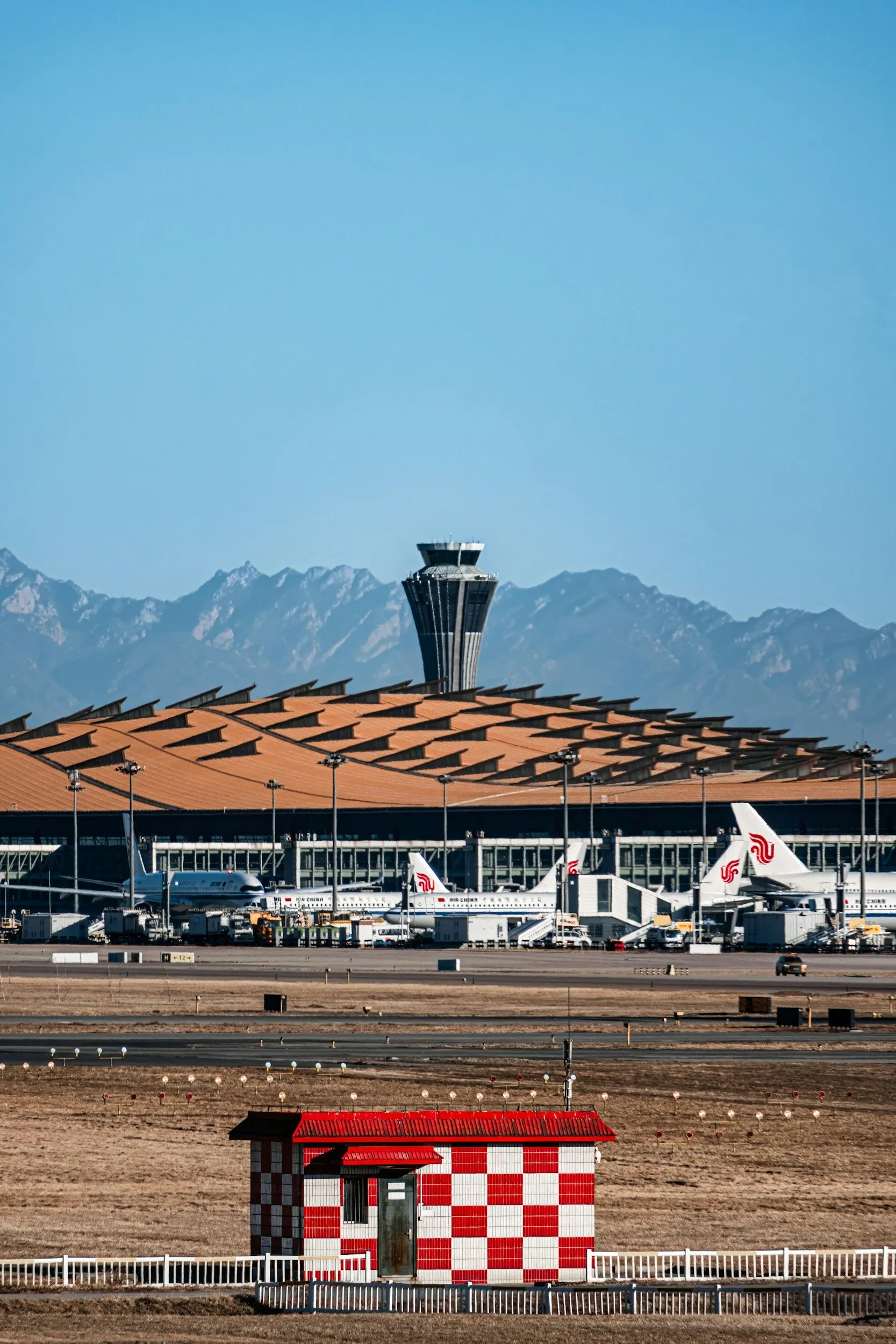 Airplane terminal with planes, control tower, mountain range in the background, a small red and white building in the foreground, and clear blue sky.