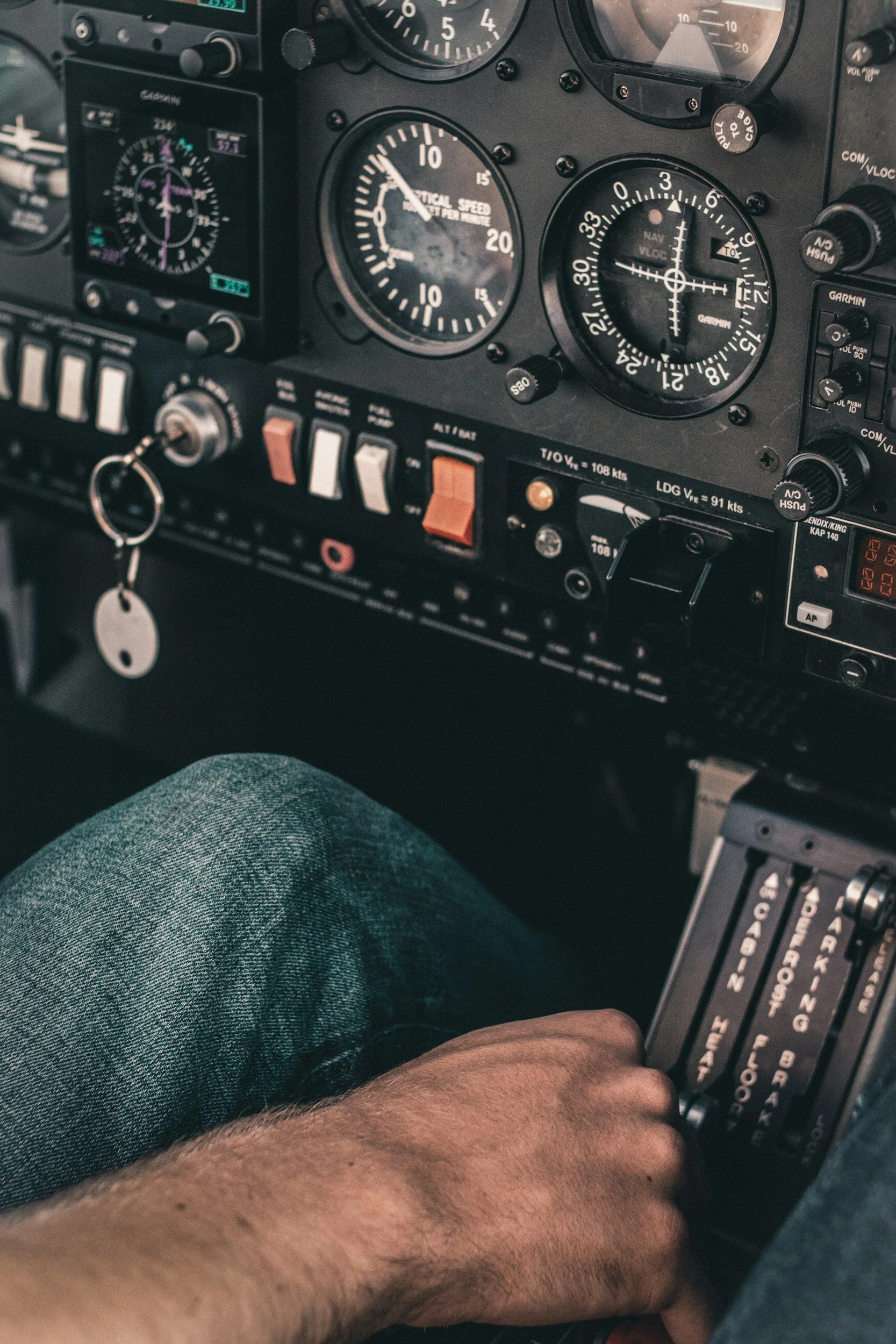 Close-up of an airplane cockpit instrument panel with gauges and controls, and a person's arm and leg visible.