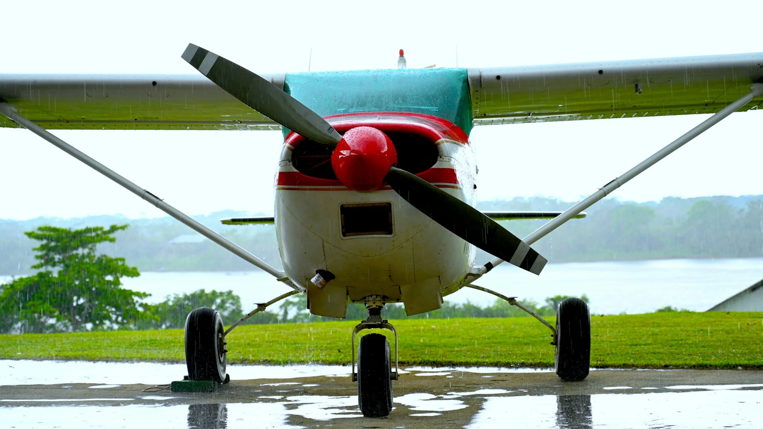 Small airplane parked outdoors in the rain with water on the ground, green landscape and river in the background.