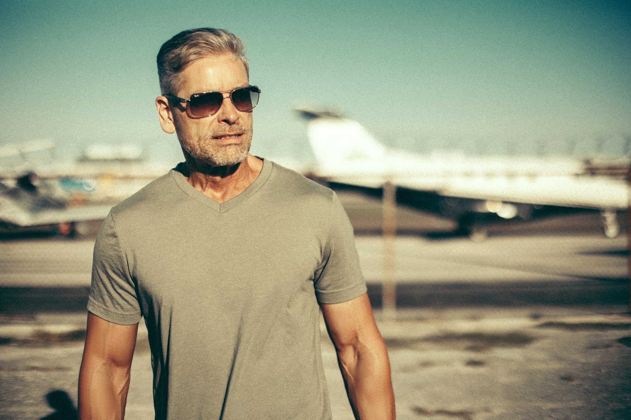 A middle-aged man with gray hair and a beard, wearing sunglasses and a light gray t-shirt, standing outdoors near an airport with airplanes in the background.