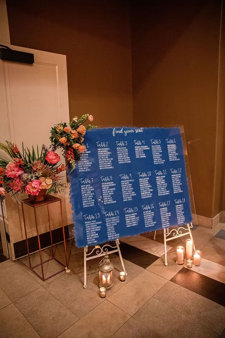 Blue wedding seating chart displaying tables 1–18 beside colorful floral arrangements and candles.