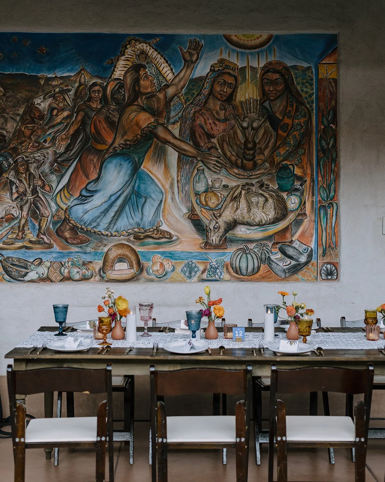 Dining room with a long table set beneath a colorful mural depicting Indigenous people, food, and animals