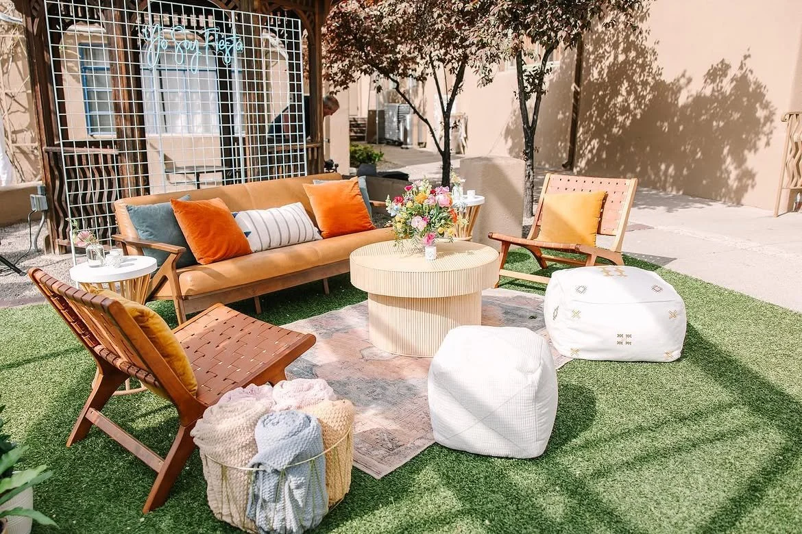 Sunlit outdoor patio lounge with a leather sofa, armchairs, poufs, coffee table, flowers, and pillows on artificial grass