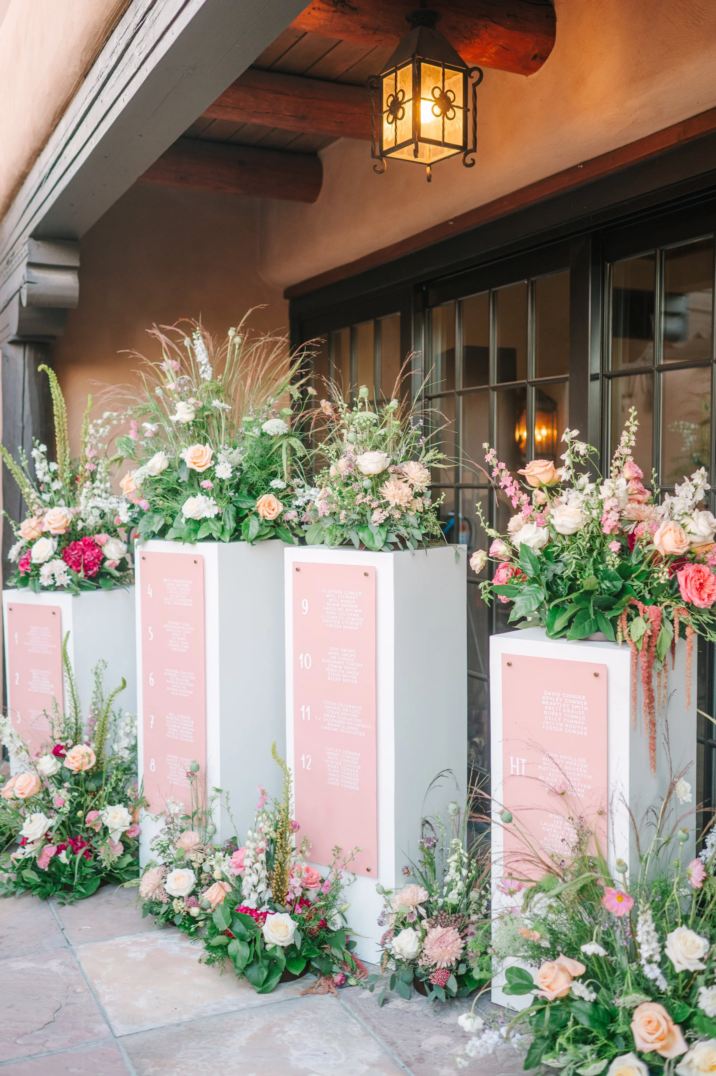 Colorful floral wedding seating display with roses, greenery, numbered pink guest cards, and a hanging lantern outside a venue