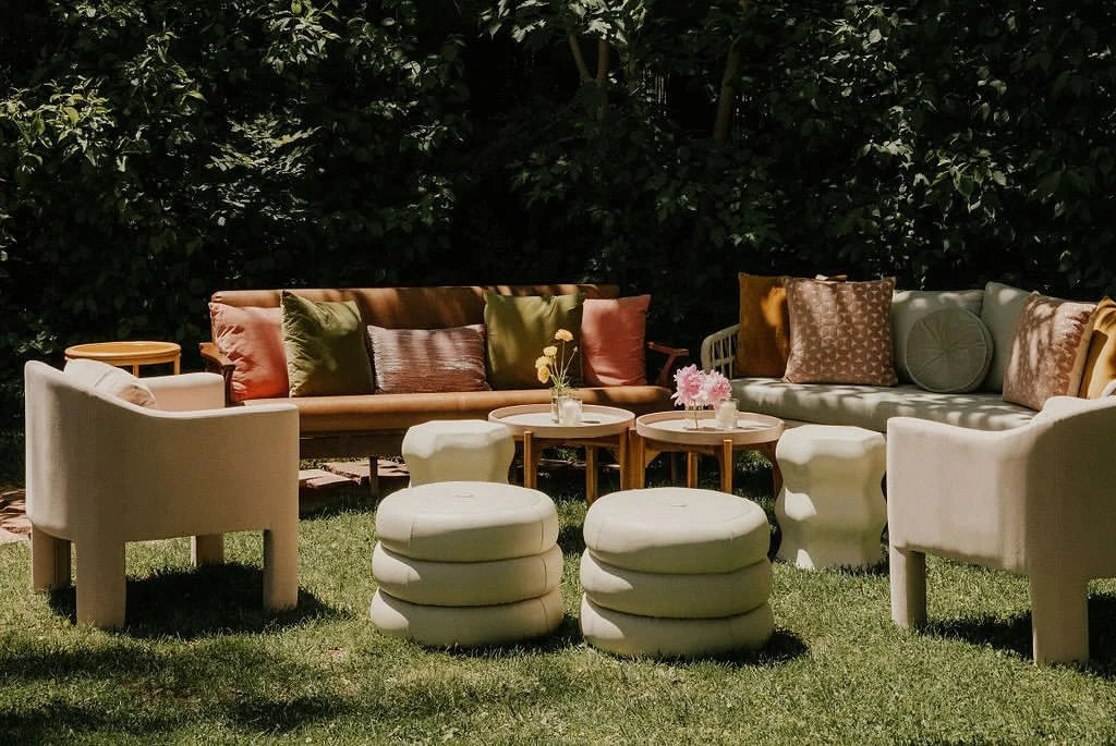 Outdoor garden lounge with cream armchairs, cushioned ottomans, wooden sofas, coffee tables, and colorful pillows