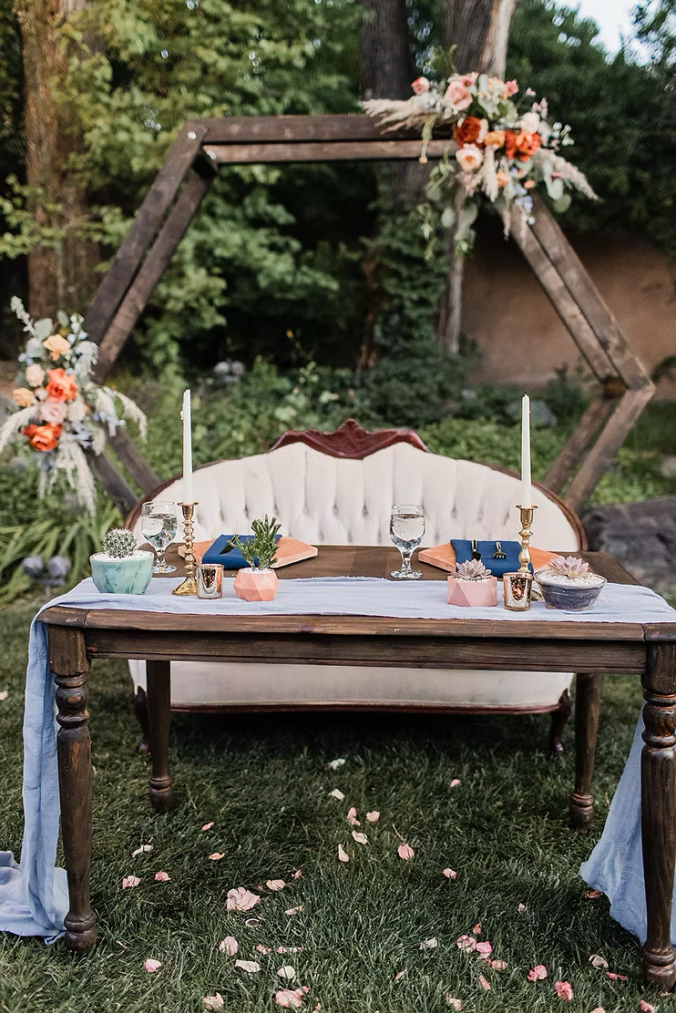 Outdoor wedding sweetheart table with floral arch, tufted loveseat, candles, place settings, and succulents