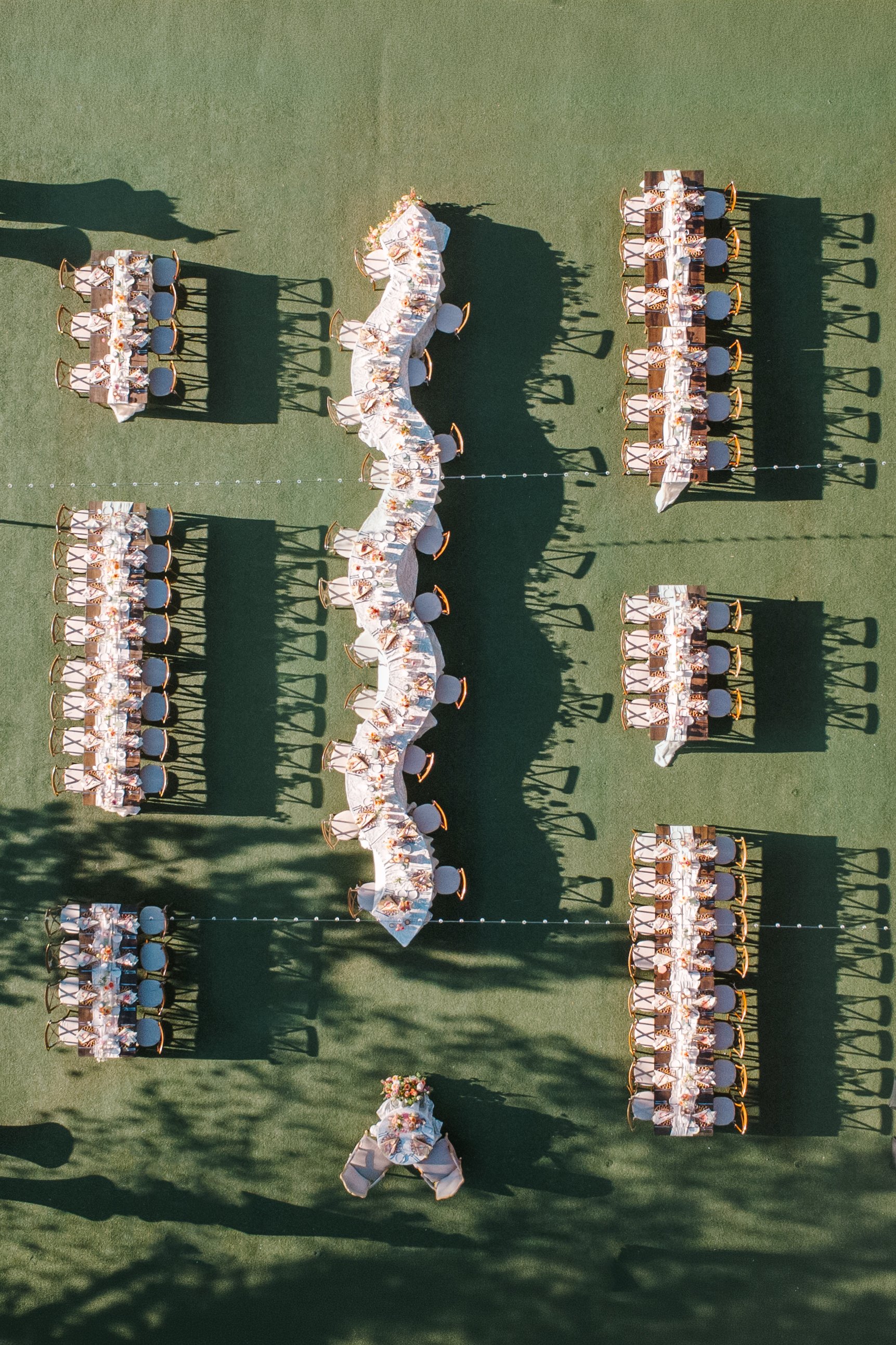 Aerial view of long outdoor banquet tables arranged on green grass, decorated with white linens, flowers, place settings, and chairs.