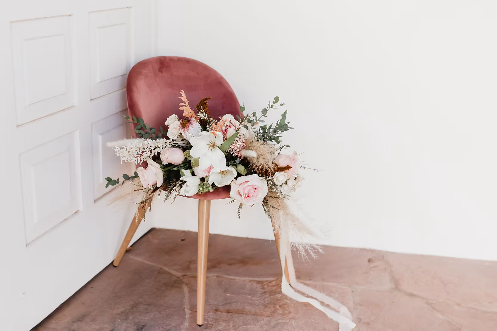 Blush velvet chair decorated with a romantic floral arrangement of roses, white flowers, eucalyptus, and dried botanicals