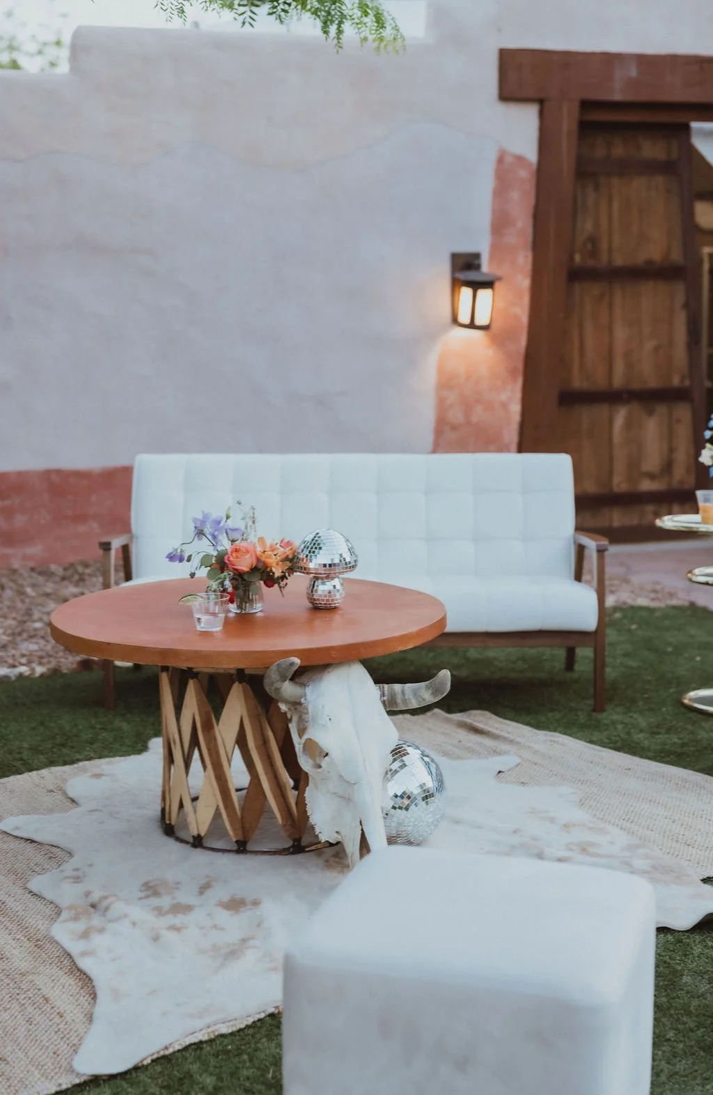 Boho outdoor lounge with a white sofa, round wooden table, floral centerpiece, disco balls, and bull skull decor.