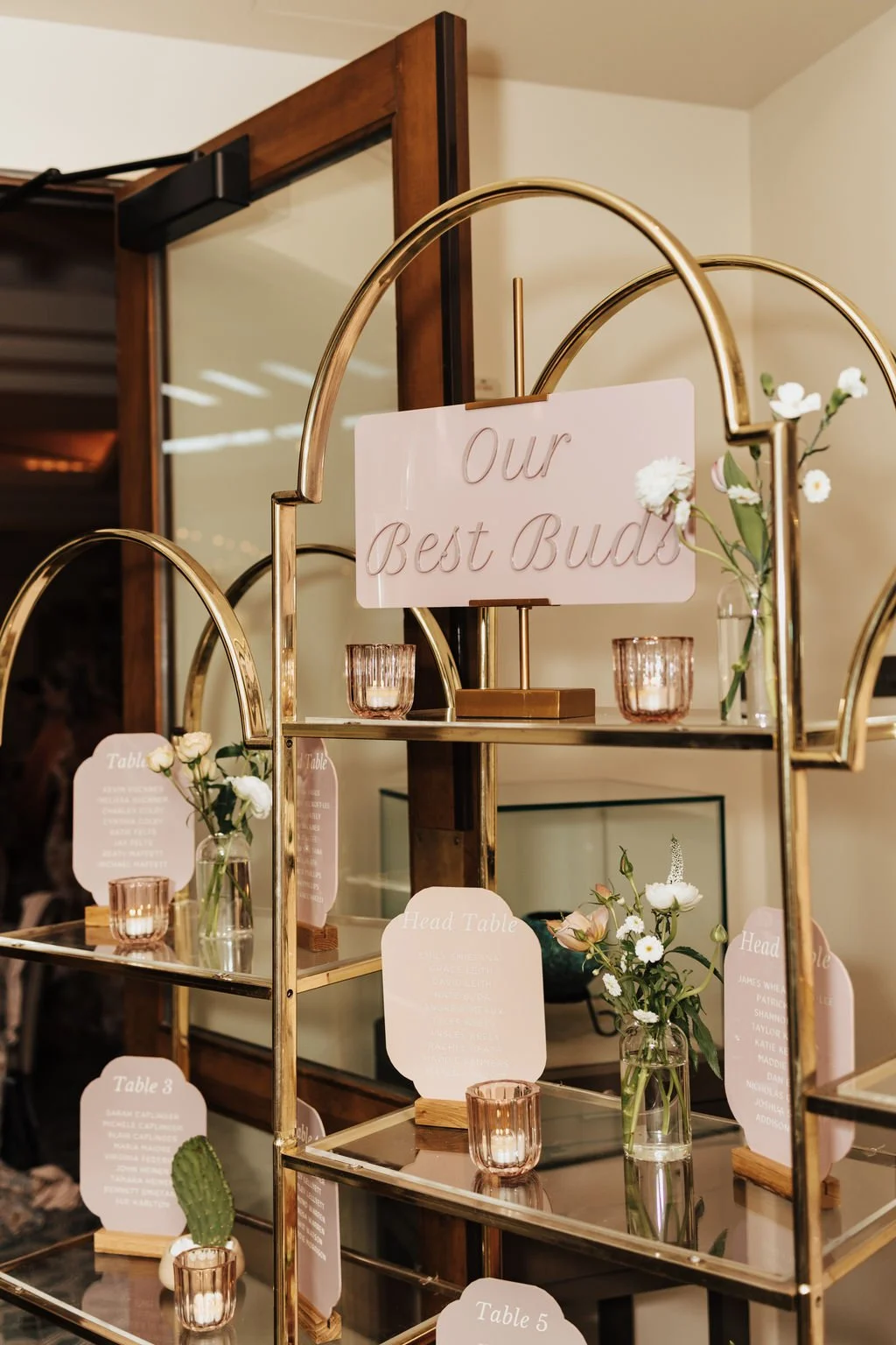 Wedding reception seating chart display with a mirrored gold shelving unit, blush table cards, flowers, candles, and a central “Our Best Buds” sign