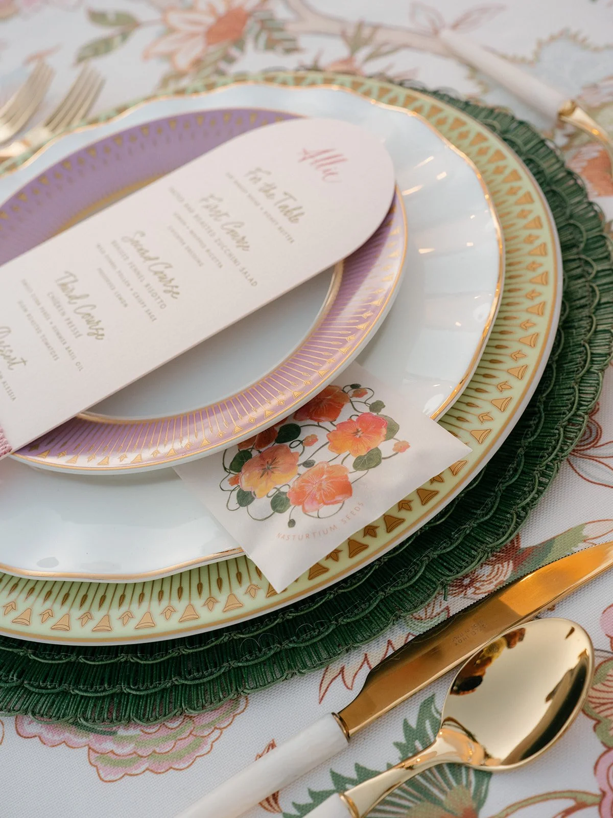 Elegant dinner table setting with layered plates, gold utensils, a printed menu with floral design, placed on a floral tablecloth.