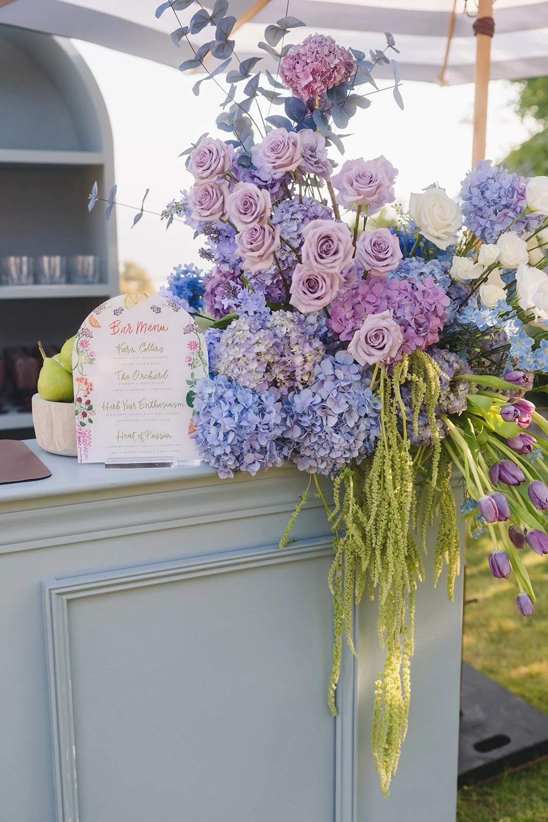 A flower arrangement with purple and white roses, lavender, and other purple and green flowers on a white table. A bar menu card is also on the table.