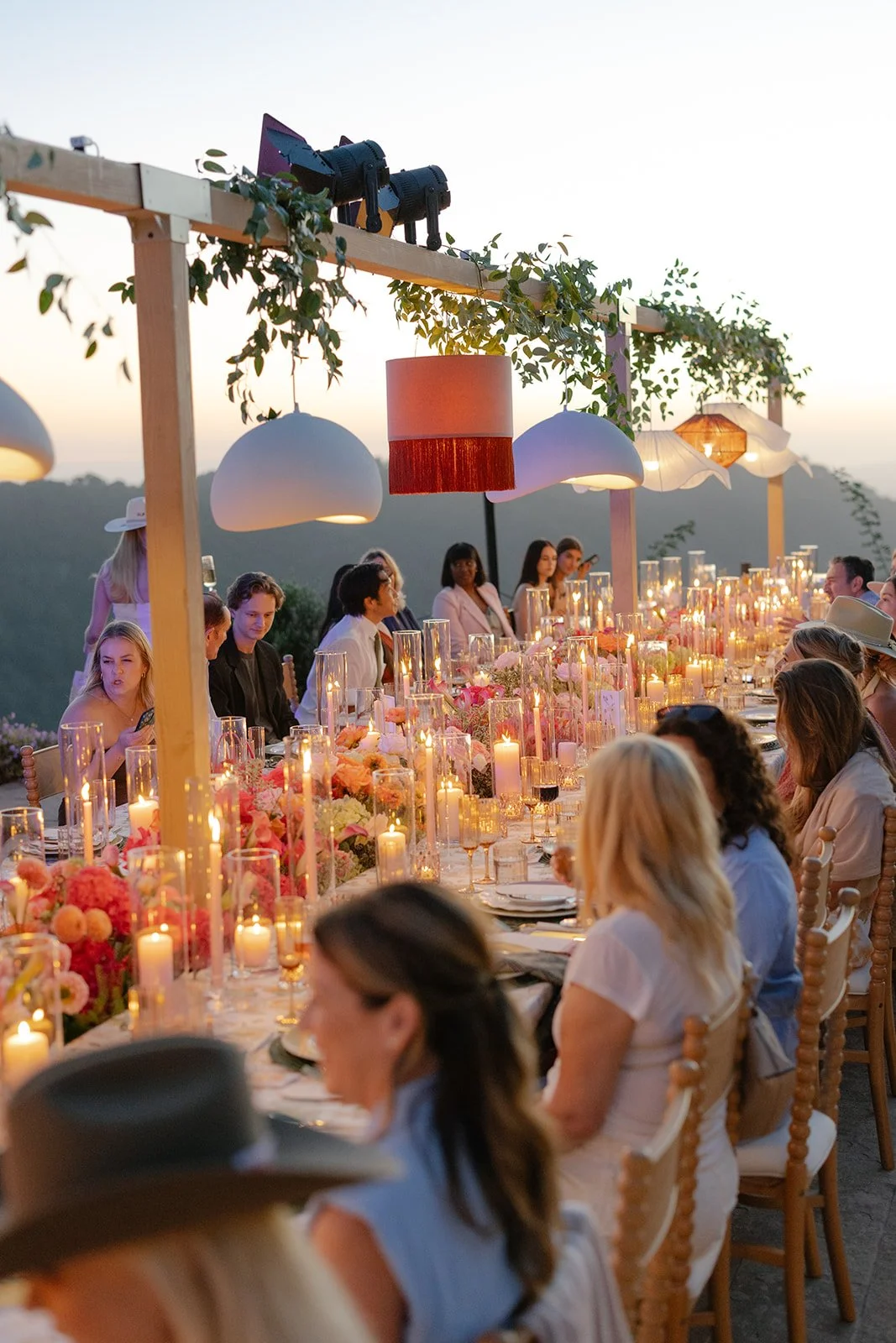 A sunset outdoor dining event with a long table decorated with candles and floral arrangements, surrounded by seated guests, with a wooden pergola overhead adorned with hanging lamps and greenery.