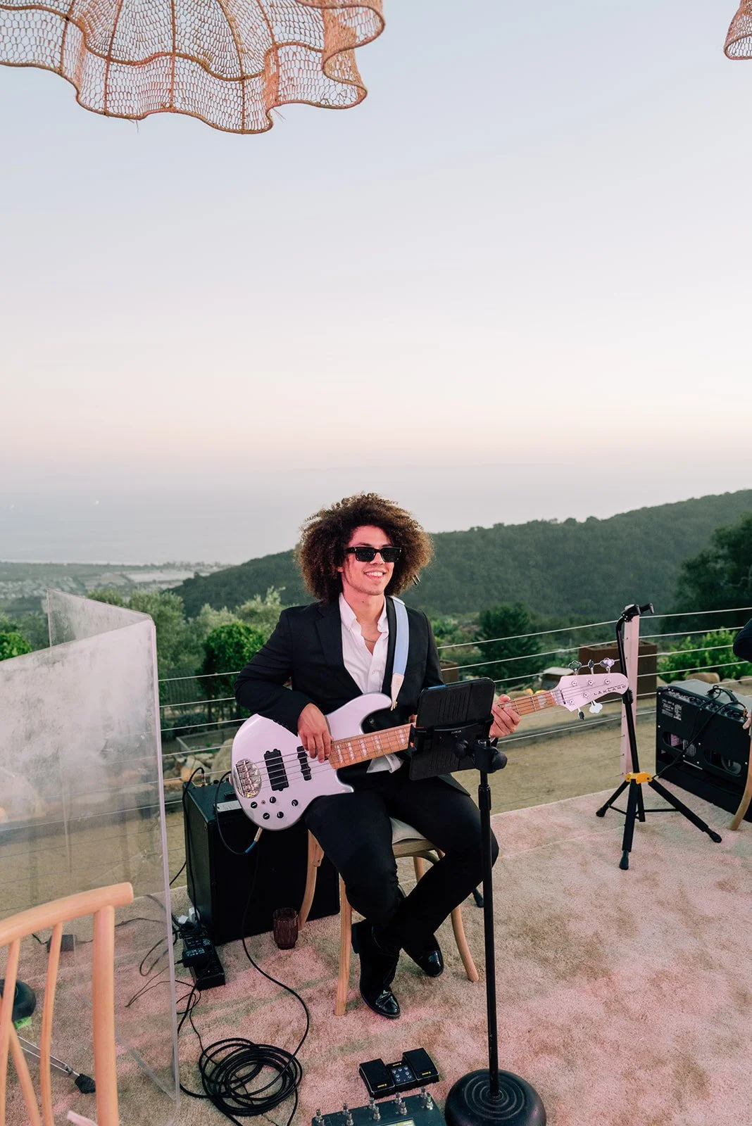 A person with curly hair, sunglasses, and a black suit jacket sitting outdoors, holding an electric bass guitar, with a scenic view of hills and sky in the background.