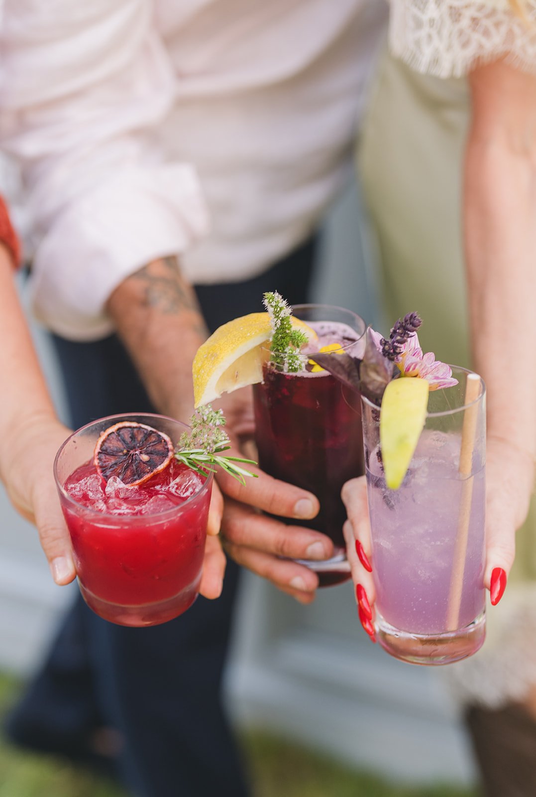 Three people holding colorful drinks with garnishes, including lemon slices, herbs, and edible flowers, outdoors.