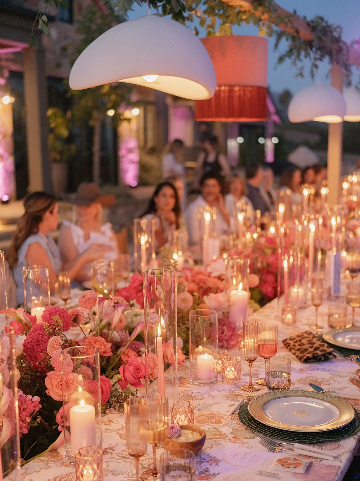 An outdoor dinner party at dusk with a long table decorated with pink flowers and lit candles, surrounded by people sitting and chatting under large white and pink lamps hanging from above.
