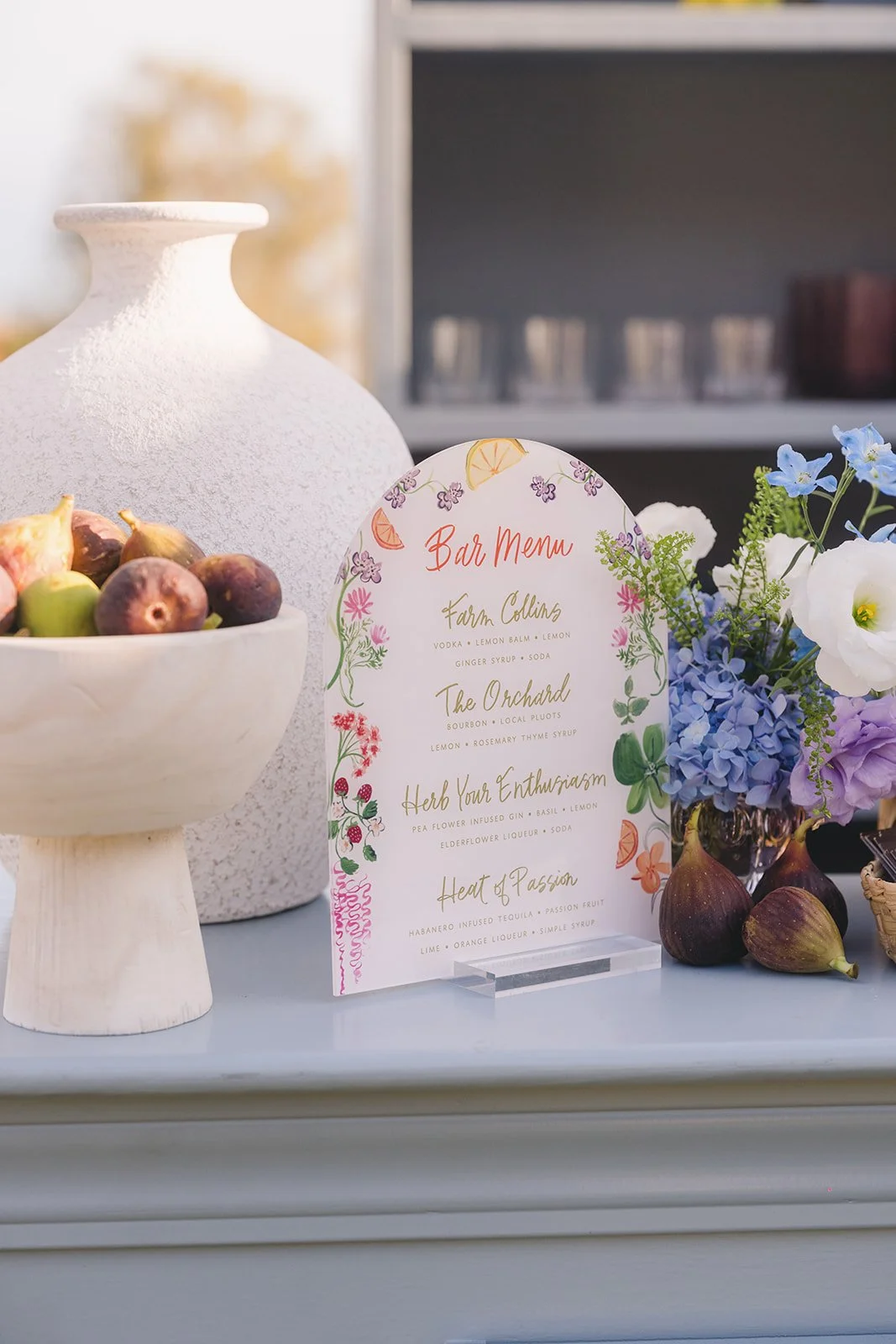A bar menu card displayed on a table surrounded by flowers, figs, and a white vase, with a blurred background.