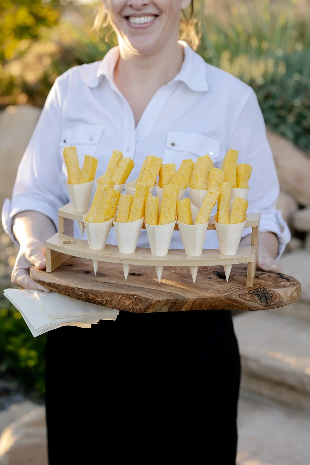 Person holding a wooden tray with several cups of French fries in paper cones, outdoors with natural background.