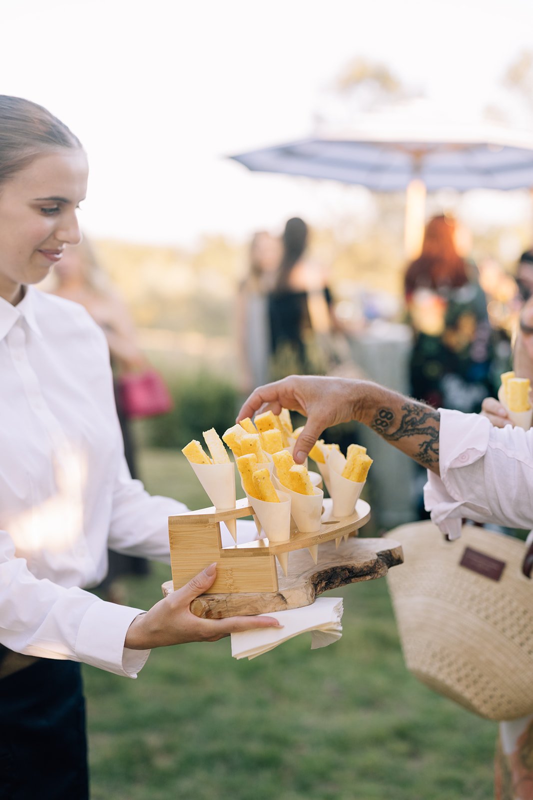 A woman in a white shirt receiving a tray of what appears to be bread or cheese sticks in an outdoor setting with people and umbrellas in the background.