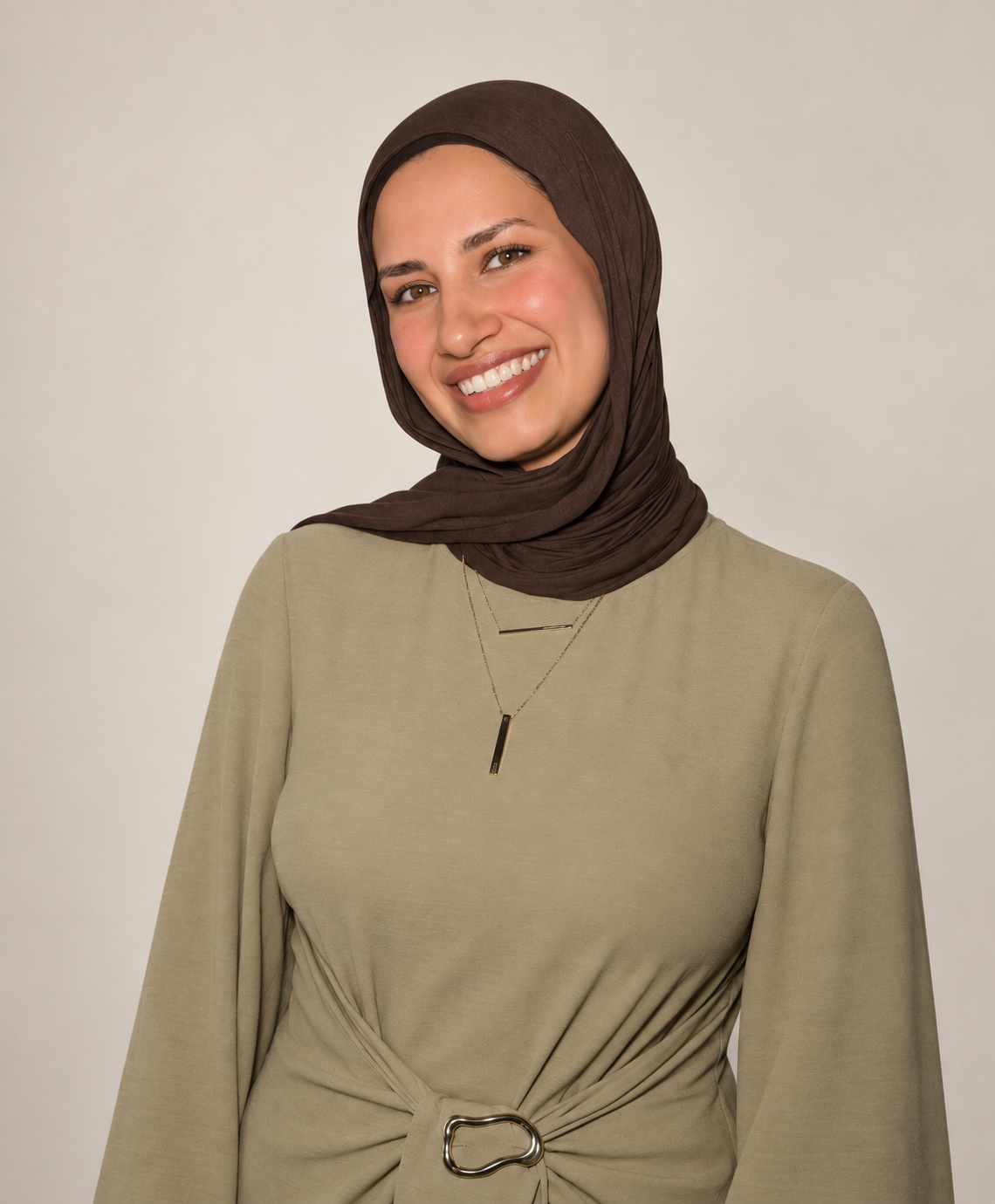 Junge Frau mit braunem Kopftuch, lächelnd, trägt beige Kleid mit Gürtel und Schmuck