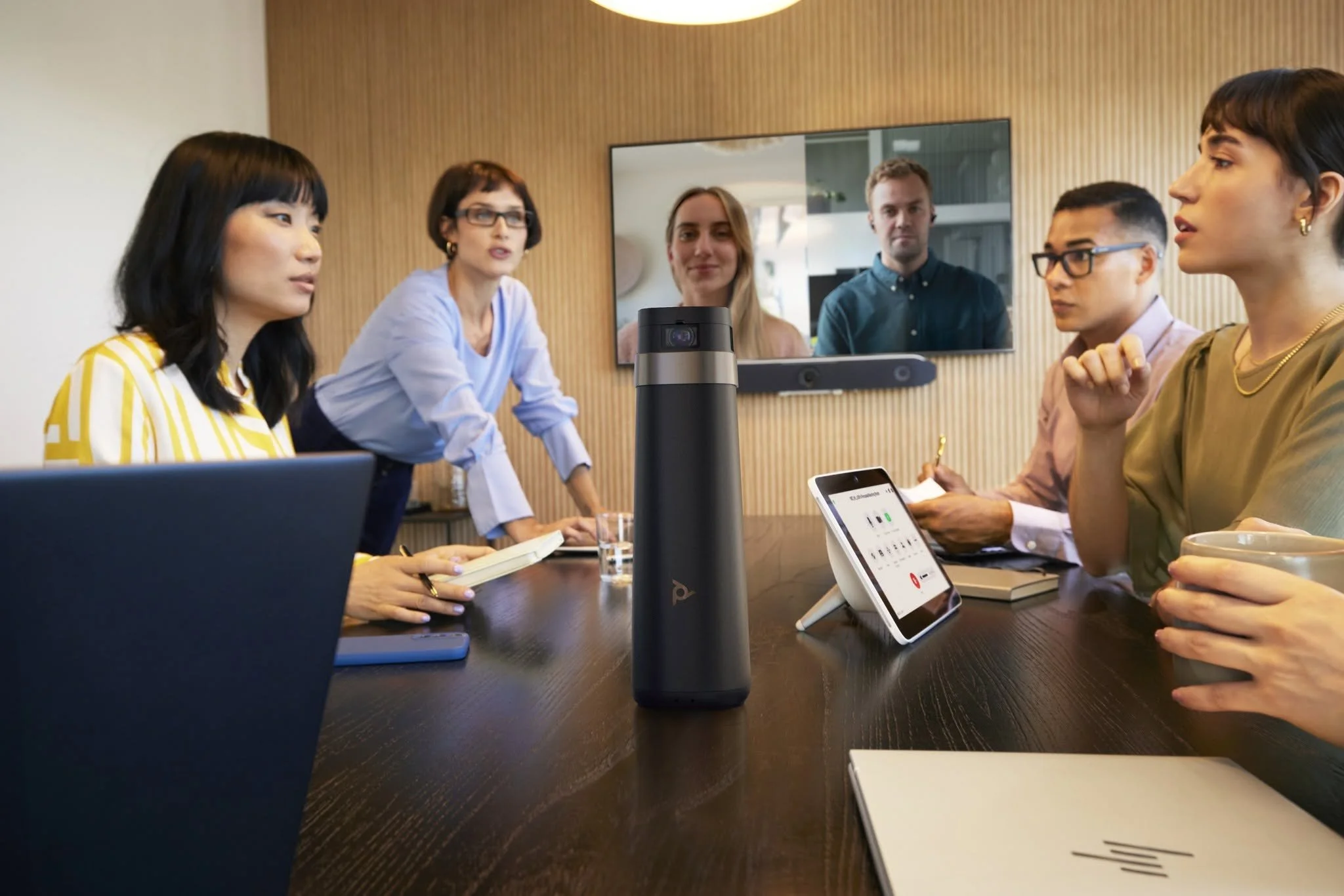A group of six diverse professionals in a meeting room, with a large screen showing a video call, a conference phone on the table, and documents, engaged in a discussion.