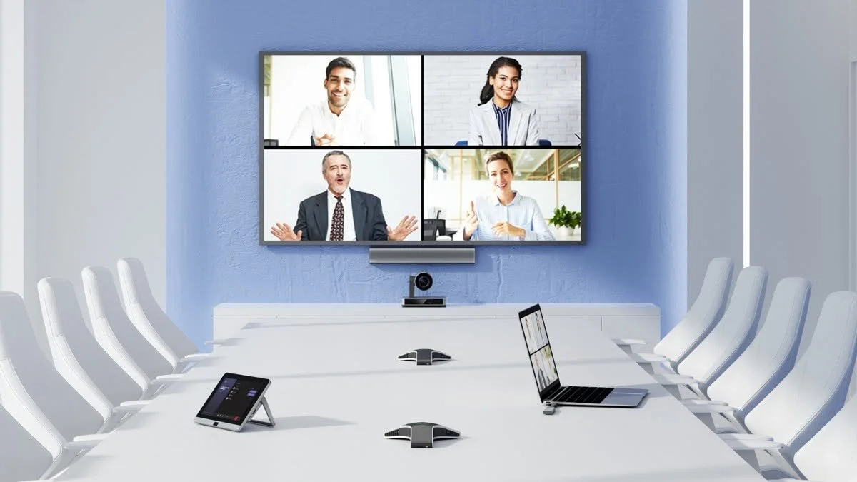 A modern conference room with a large screen displaying a video call with four people, surrounded by white chairs, microphones, a laptop, and a tablet.