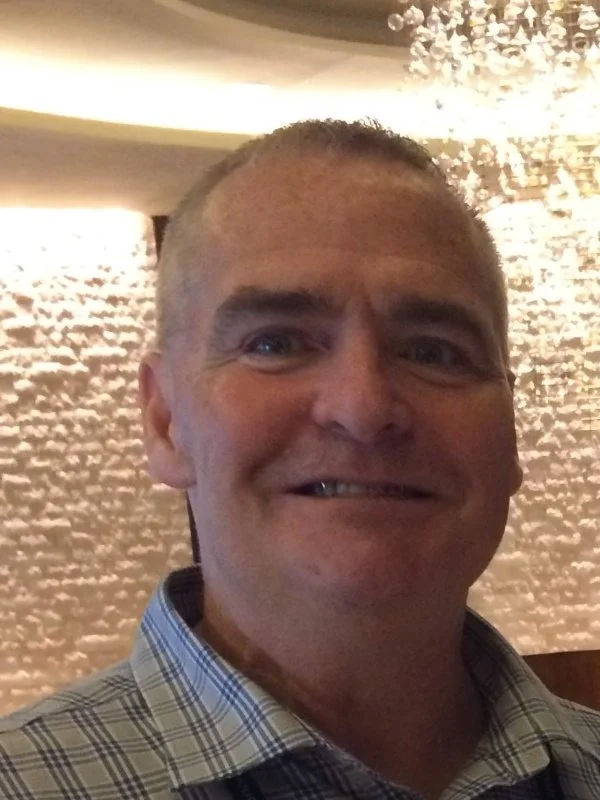 A smiling man taking a selfie indoors with textured wall and chandelier in the background.
