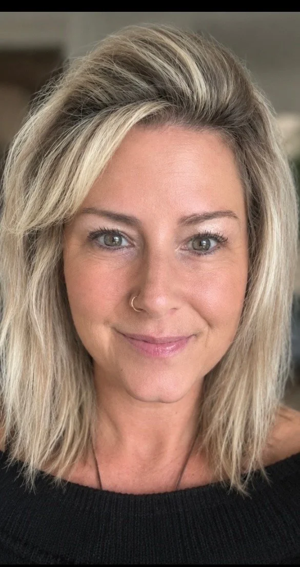 Close-up of a woman with blonde, shoulder-length hair, light skin, and a small nose ring, smiling at the camera.