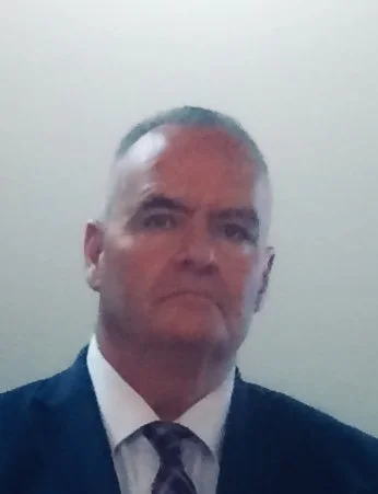 A middle-aged man with short gray hair and a serious expression, wearing a dark suit, white shirt, and striped tie, standing against a plain light-colored background.