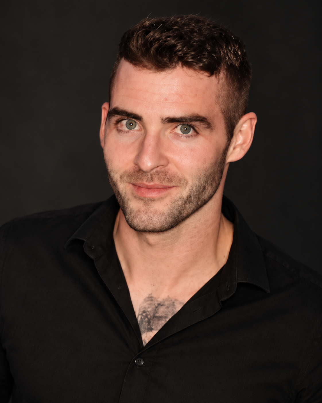 Headshot of a young man with short, dark, curly hair and gray eyes, wearing a black shirt, smiling slightly against a dark background.