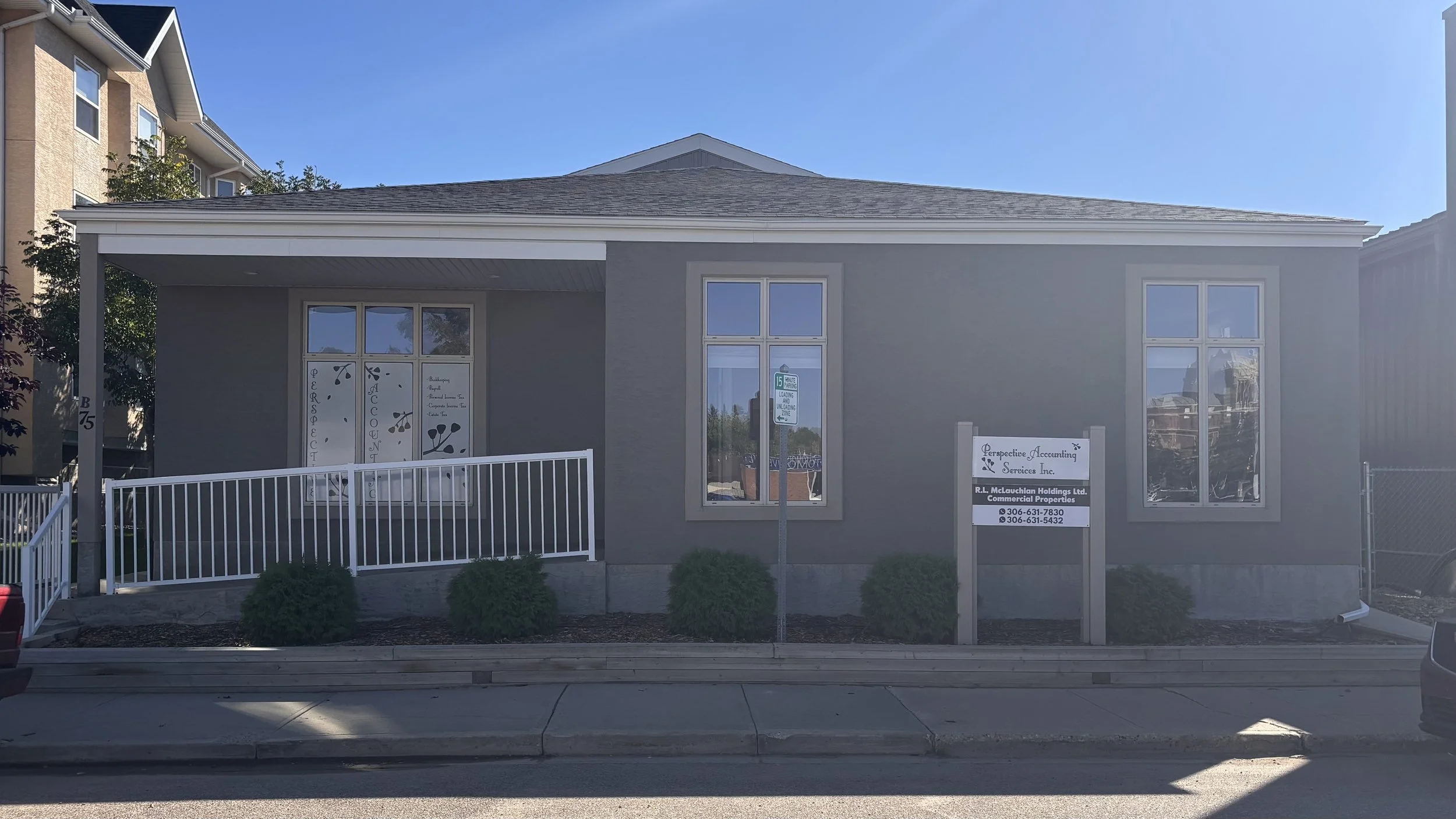 A small commercial building with a sign for Perspective Accounting Services Inc., located on a city street. The building has gray walls, four windows with decorative decals, a white fence, and some small bushes in front. There is a street sign indicating a parking zone and a sidewalk.
