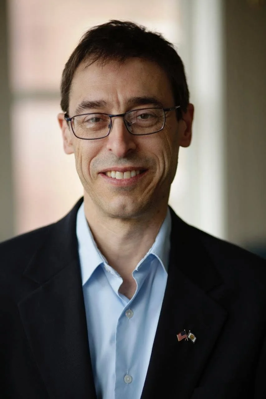 A man with short dark hair and glasses, smiling, wearing a blue shirt and a black blazer with American flag pin, standing indoors with a blurred background.