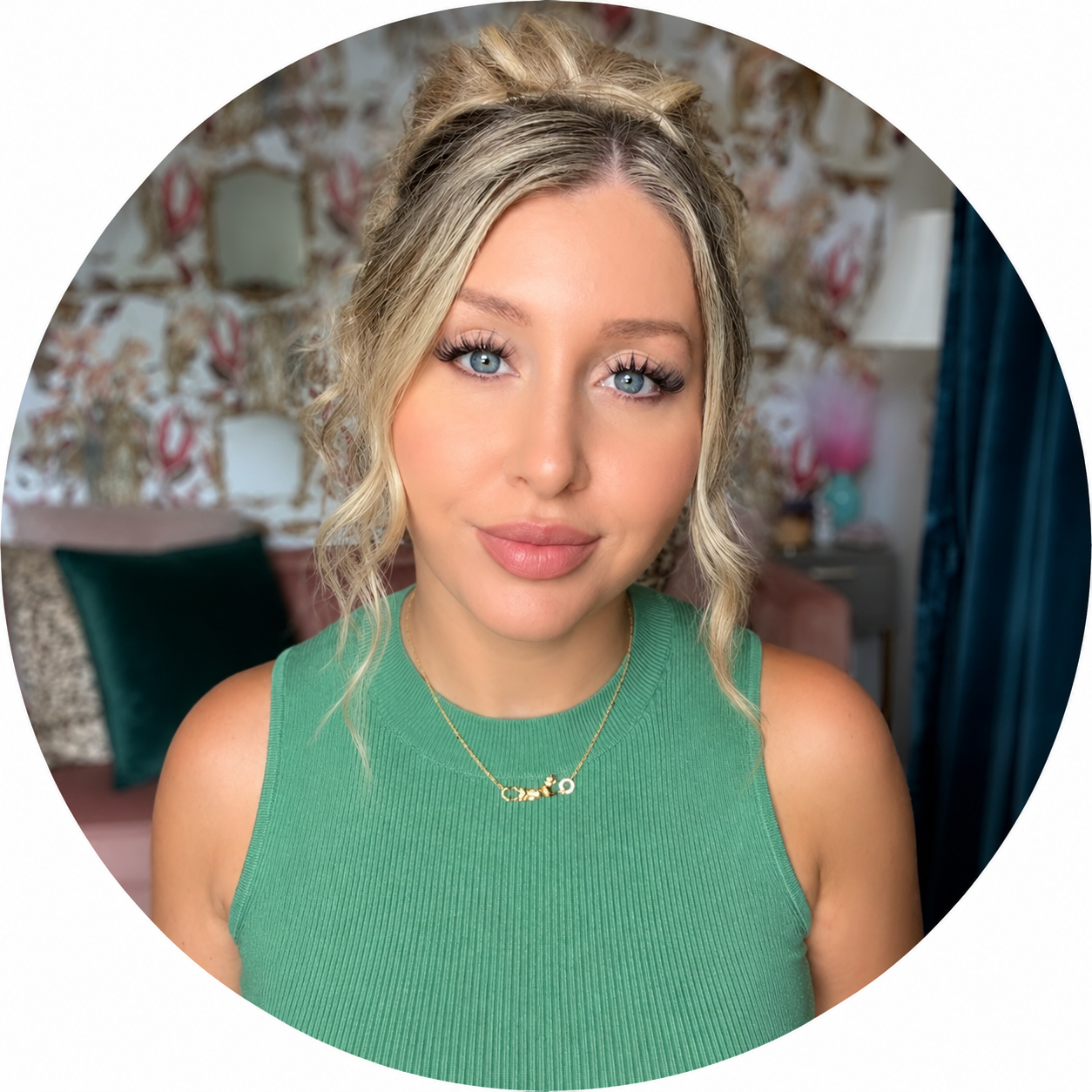 A young woman with blonde hair and blue eyes wearing a green sleeveless top and a gold necklace. She is indoors with a floral-patterned wall, pillows, and a lamp in the background.