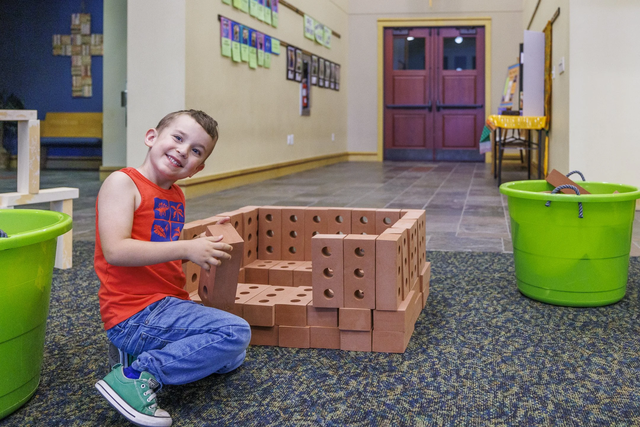 A young boy with brown hair wearing a red tank top with blue pattern, blue jeans, and teal sneakers, is sitting on the floor and smiling while building a structure with large red bricks in a classroom or play area.