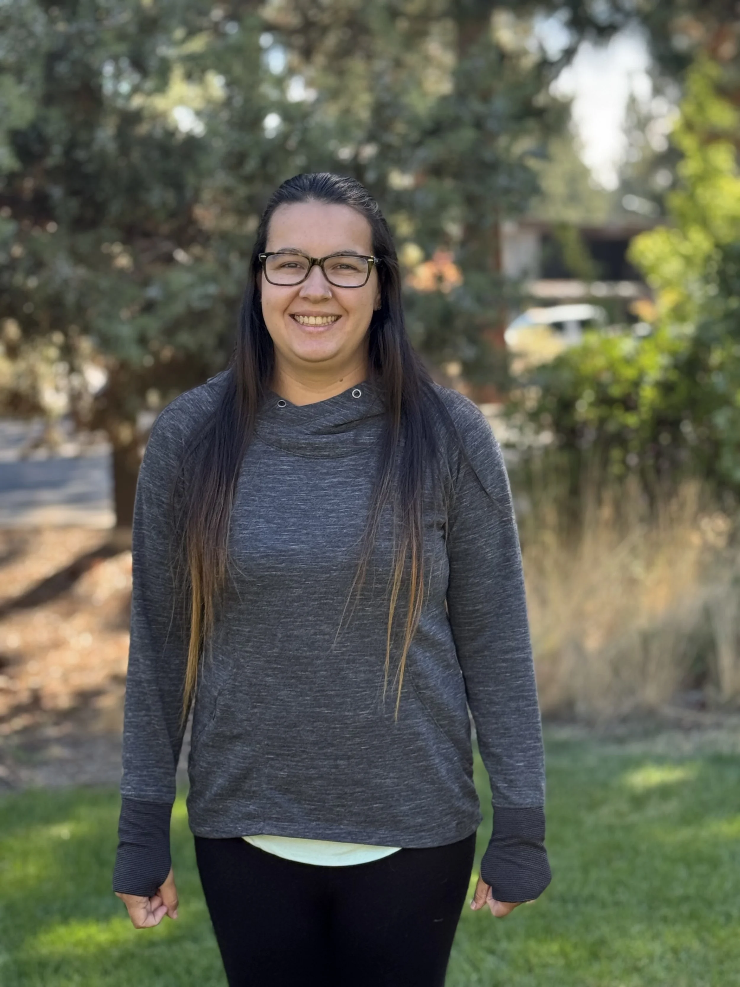 A woman with long dark hair, glasses, and a nose ring smiling outdoors during daytime, wearing a dark gray hoodie and black pants, with trees and greenery in the background.