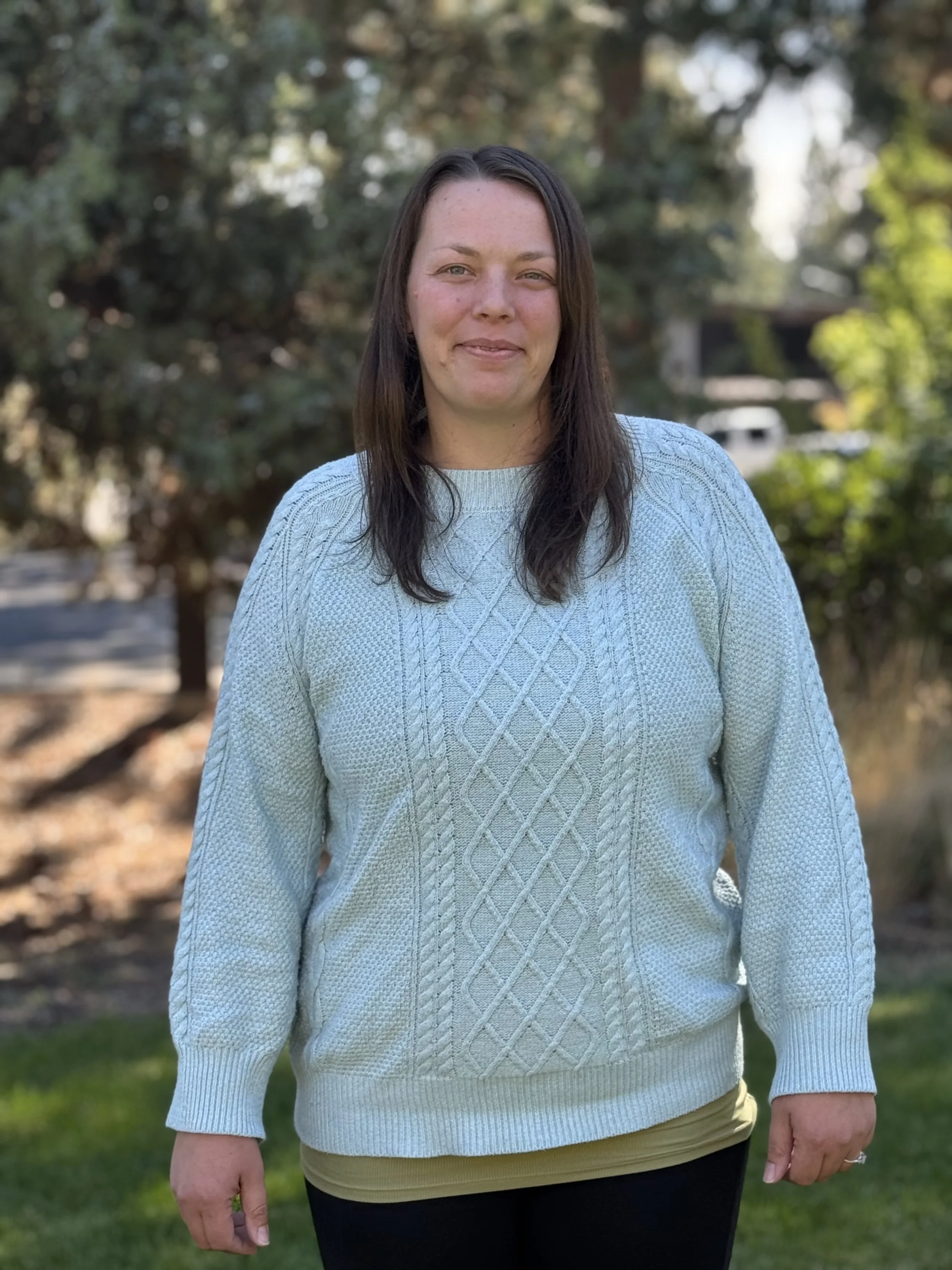 A woman with shoulder-length dark brown hair wearing a light blue cable-knit sweater and black pants standing outdoors on a sunny day with trees and a house in the background.