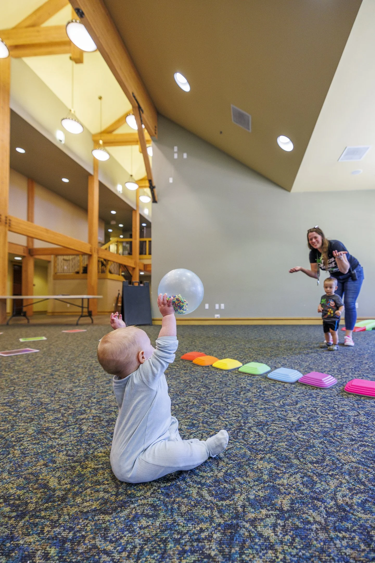 A young child with blonde hair sitting on a multicolored carpet in an indoor play area, reaching up to catch a plastic sphere filled with colorful beads. In the background, an adult woman and another child are smiling and appearing to play together, with various game equipment and colorful stepping stones on the floor.