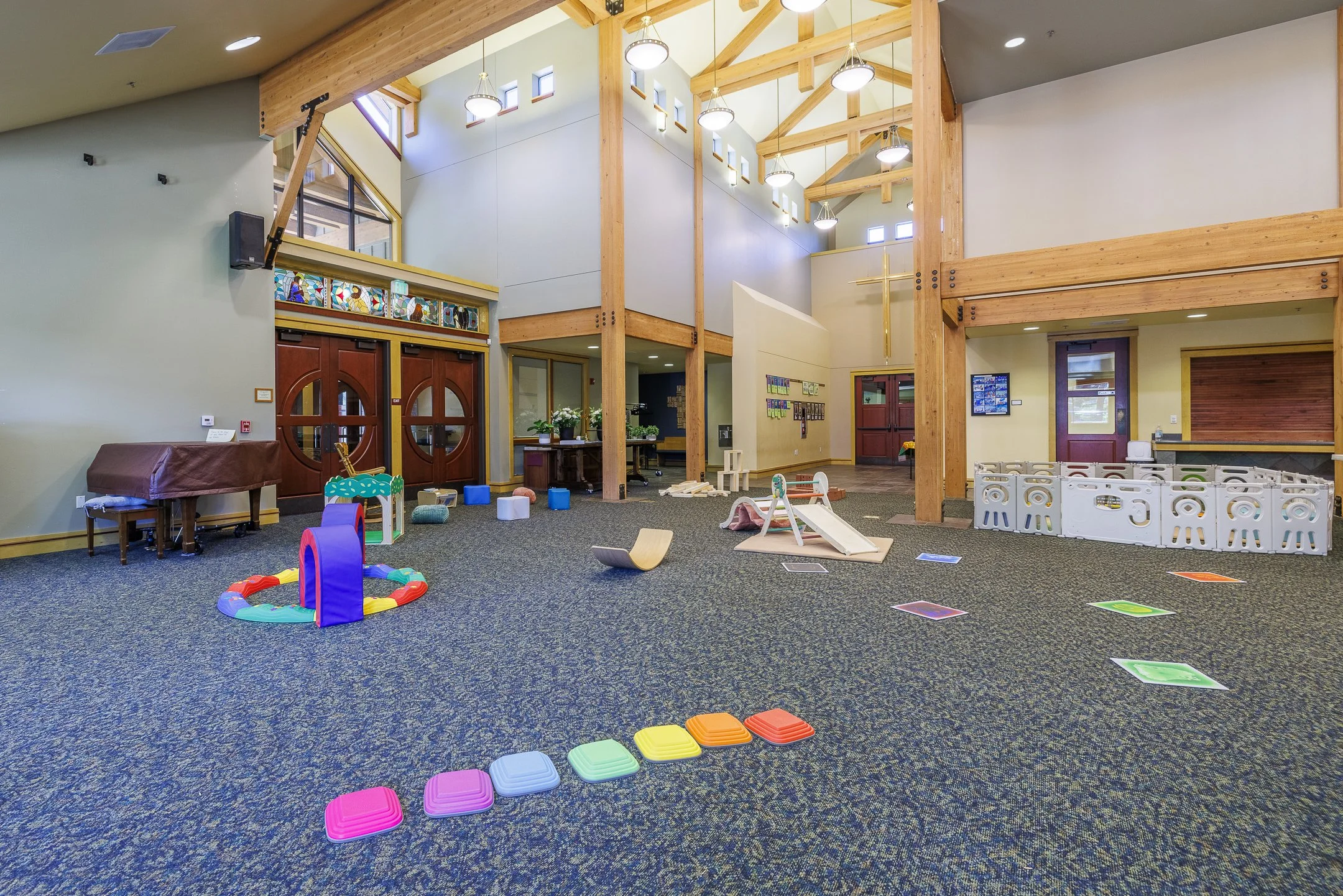 Interior of a children's playroom with colorful toys, small furniture, and a carpeted floor, featuring wooden beams and high windows.