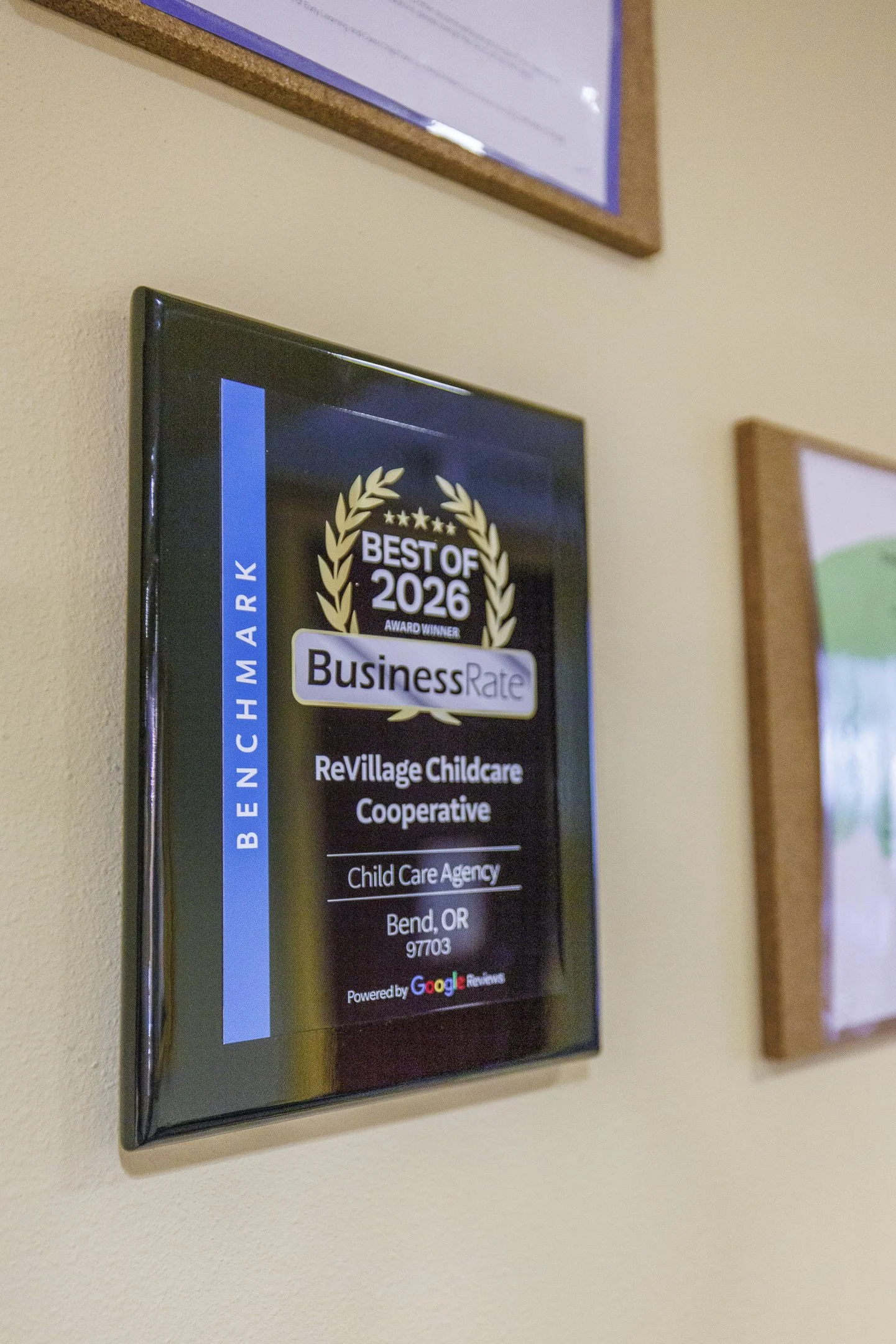 A digital display mounted on a beige wall, showing an award for being the best childcare agency of 2026, presented to ReVillage Childcare Cooperative in Bend, Oregon.