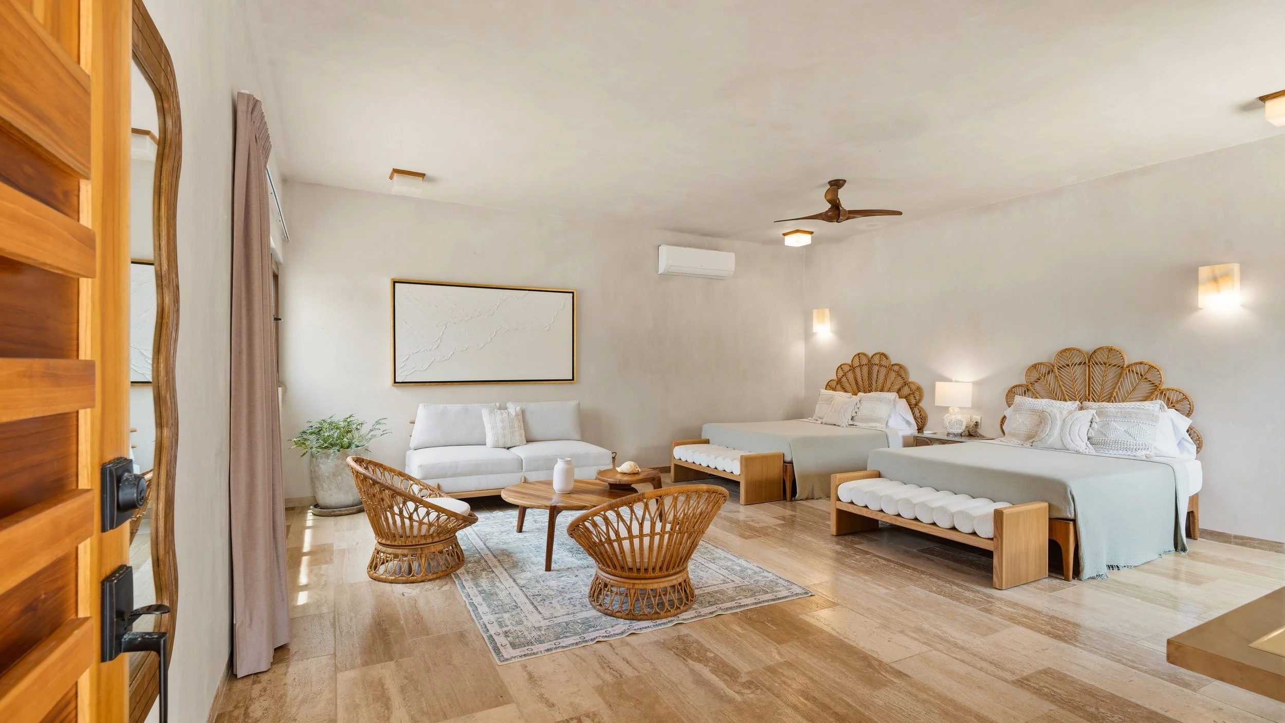 A spacious hotel room with two beds, a white sofa, wicker chairs, a wooden coffee table, and tropical decor including a large wall art and plants, with wood flooring and soft lighting.