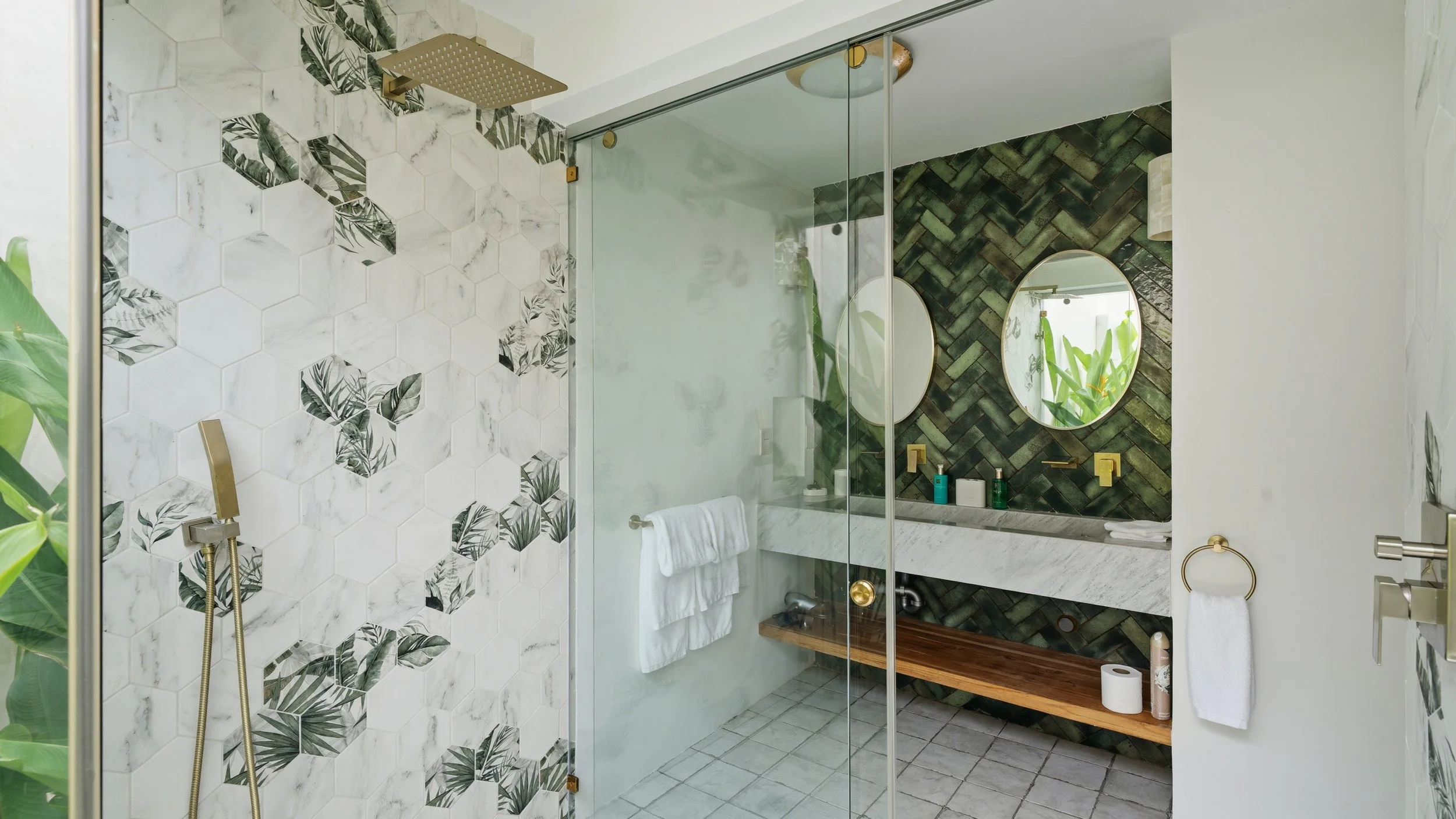 Modern bathroom with a walk-in shower, featuring white hexagonal tiles with black and green leaf patterns, and a back wall with green herringbone tiles. A large mirror and wooden bench with toiletries are also visible.
