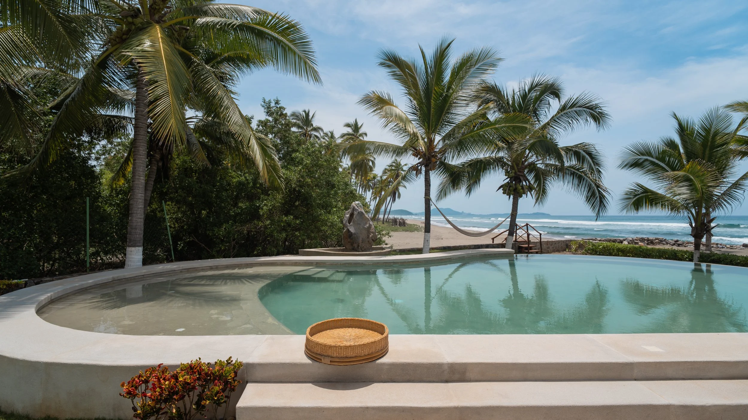 Swimming pool with a view of tropical palm trees, sandy beach, and ocean waves in the background under a blue sky.