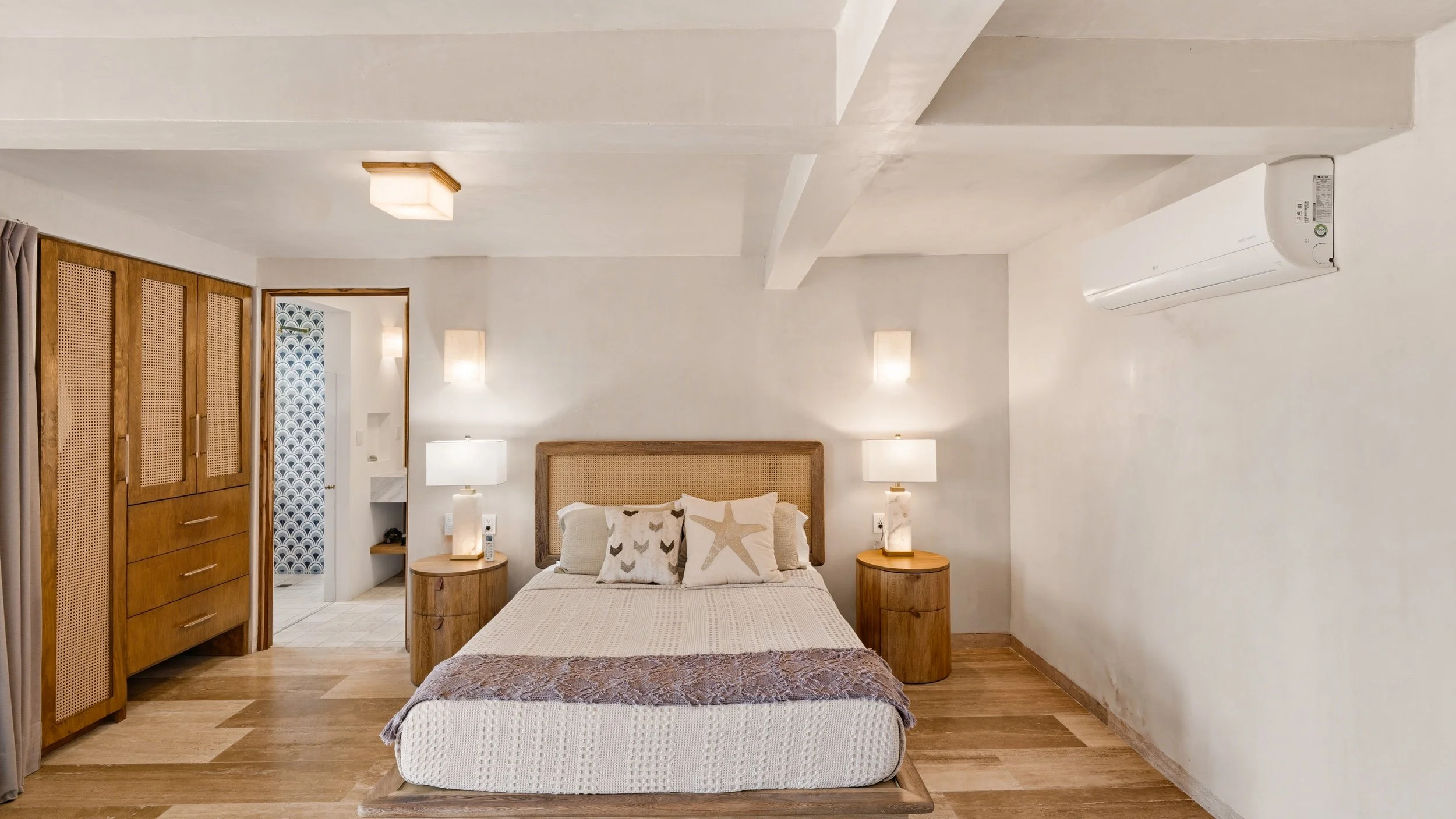 A cozy bedroom with a wooden bed frame, white bedding, and decorative pillows featuring star and fish designs. There are two wooden nightstands with lamps on each side of the bed, wooden floors, and a white wall with a split-air conditioner mounted o