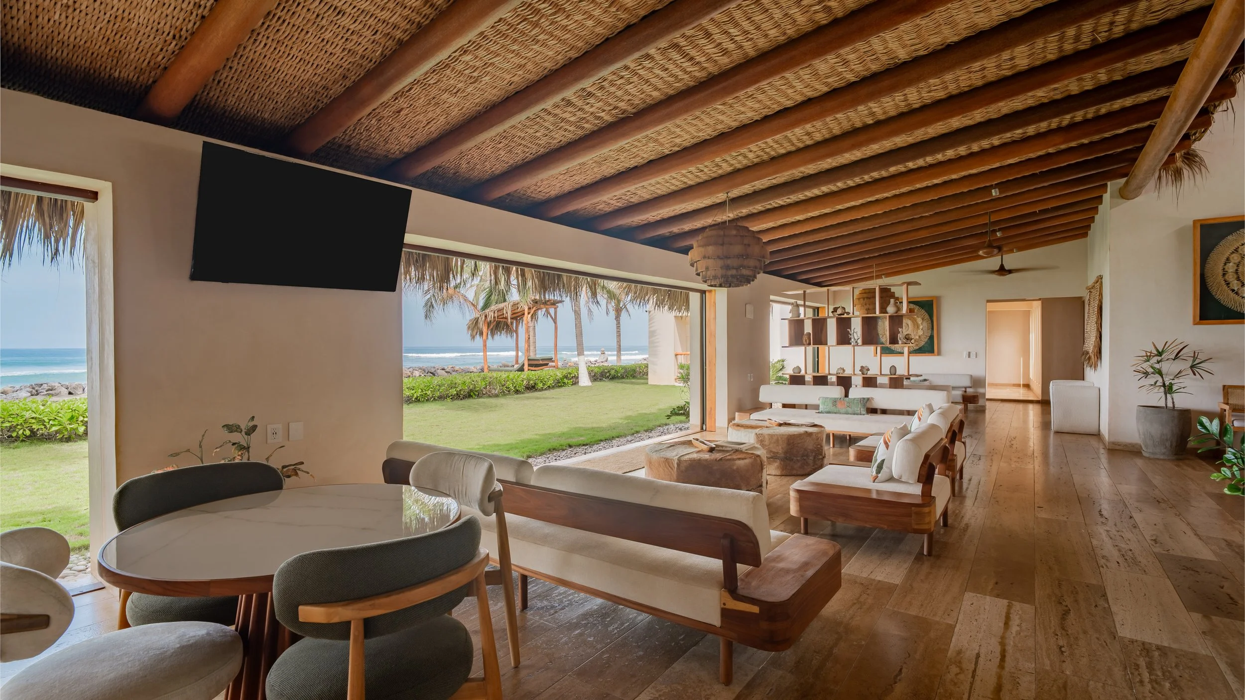 A beachfront living room with large open sliding doors showing a view of the beach, palm trees, and the ocean. The room features a wooden beamed ceiling, modern furniture, and decorative accents.