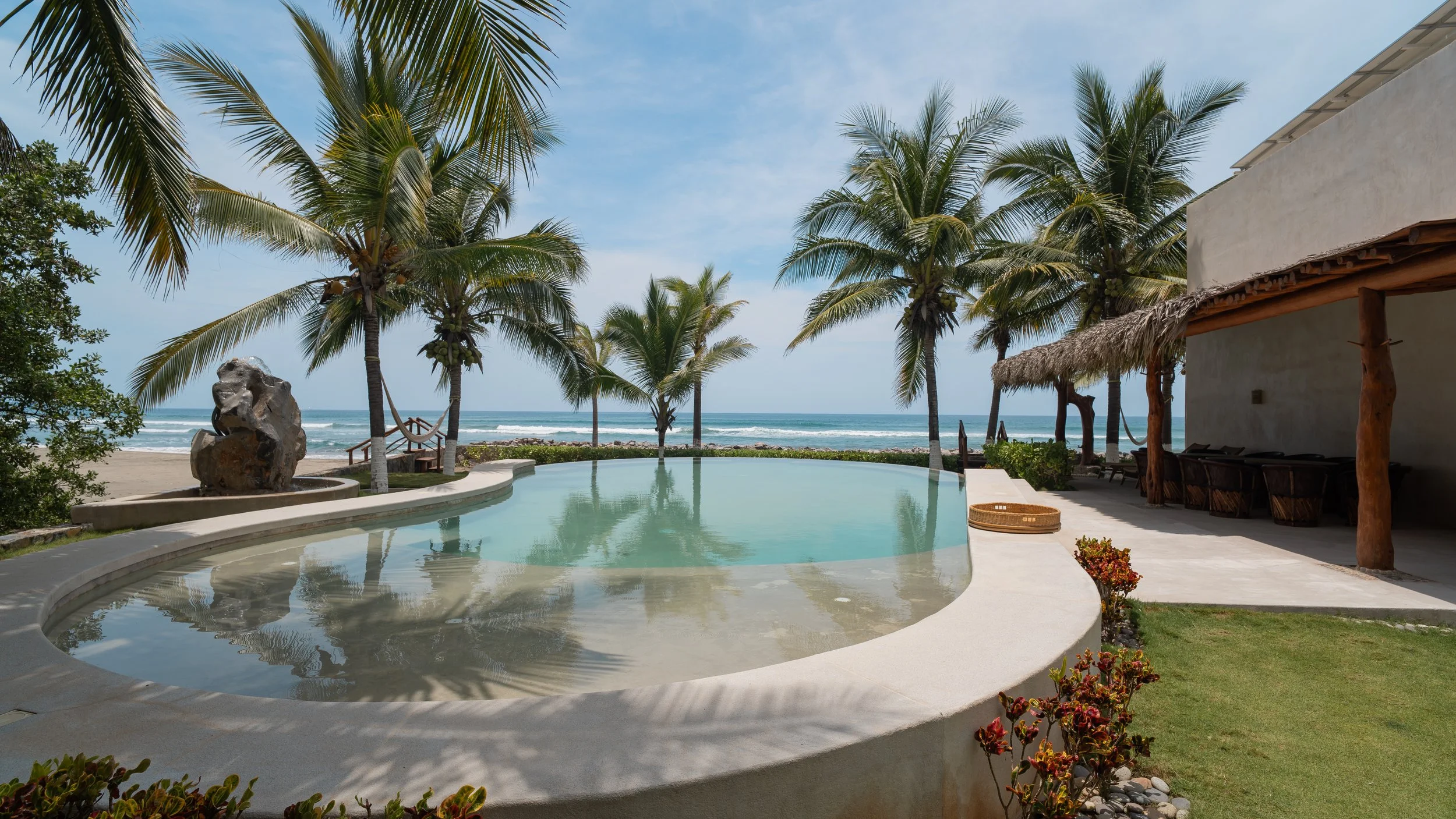 A tropical resort pool area with a view of the ocean in the background, surrounded by palm trees, thatched roof structures, lounge chairs, and decorative stones.