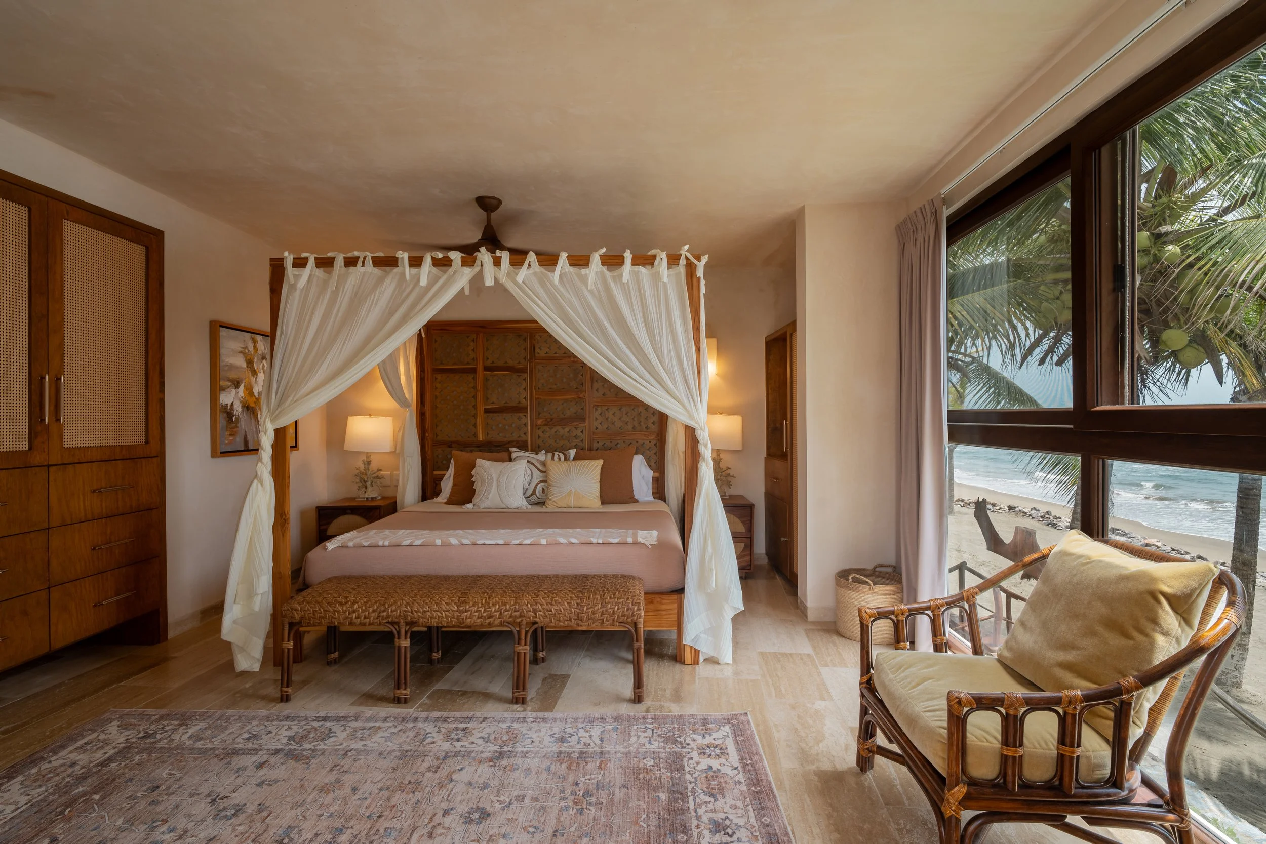 A bedroom with a canopy bed and a view of the beach and ocean through large open windows.