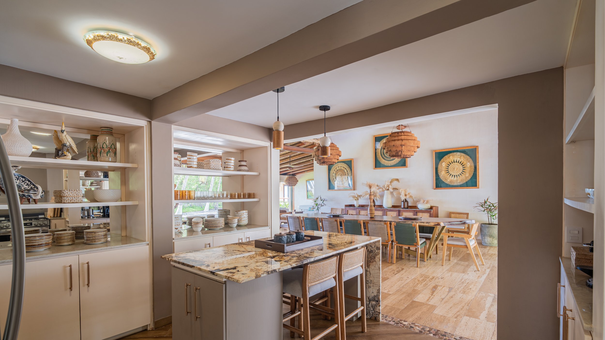 Interior view of a dining area with artwork on the wall, wooden chairs around a long table, and open shelving with dishes and decor, illuminated by natural light.